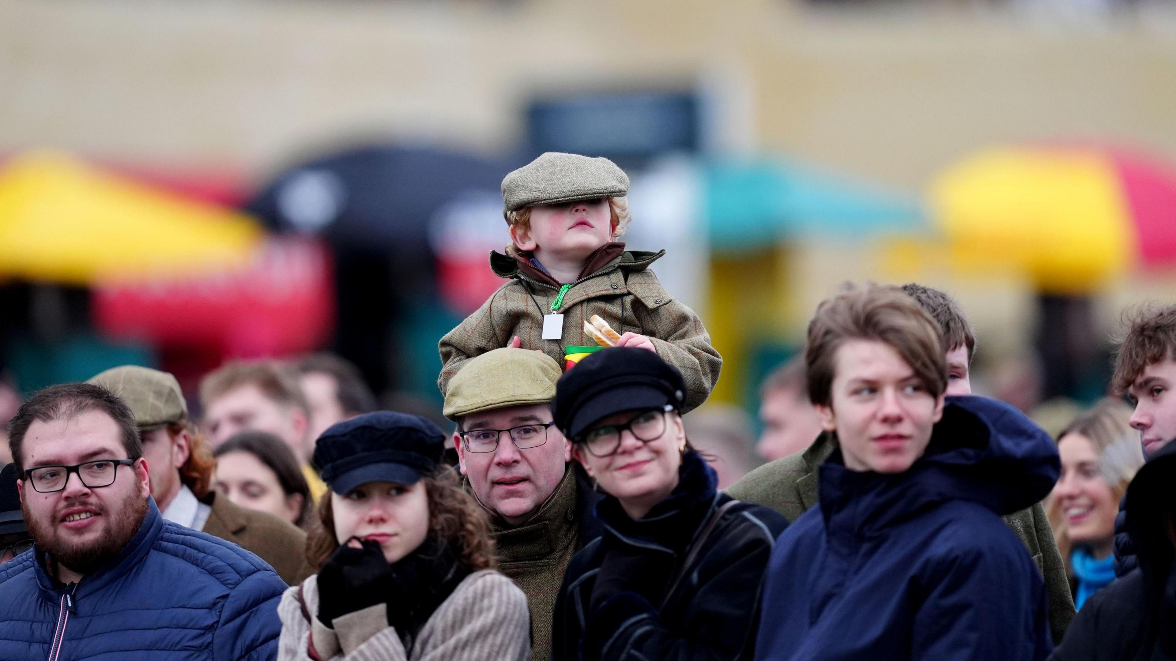 Racegoers watch the action at Cheltenham Race Course on 1 January 2025. In the picture is a young boy wearing a cap that is too large for him. He is sitting on someone else's shoulders