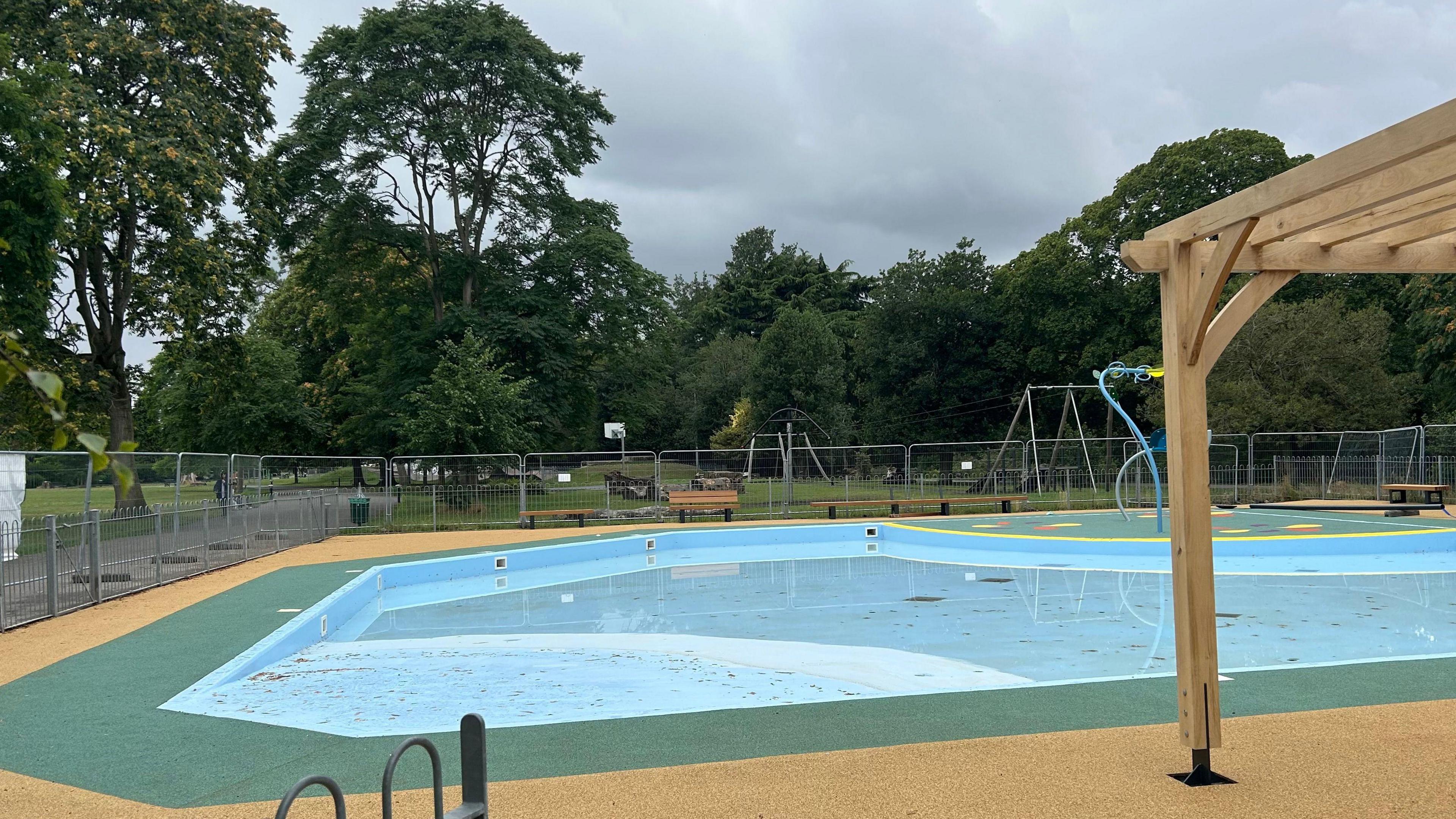 An outdoor municipal paddling pool, with a light blue base, is surrounded by a green and yellow safety surface. There is fencing around the perimeter of the pool area.