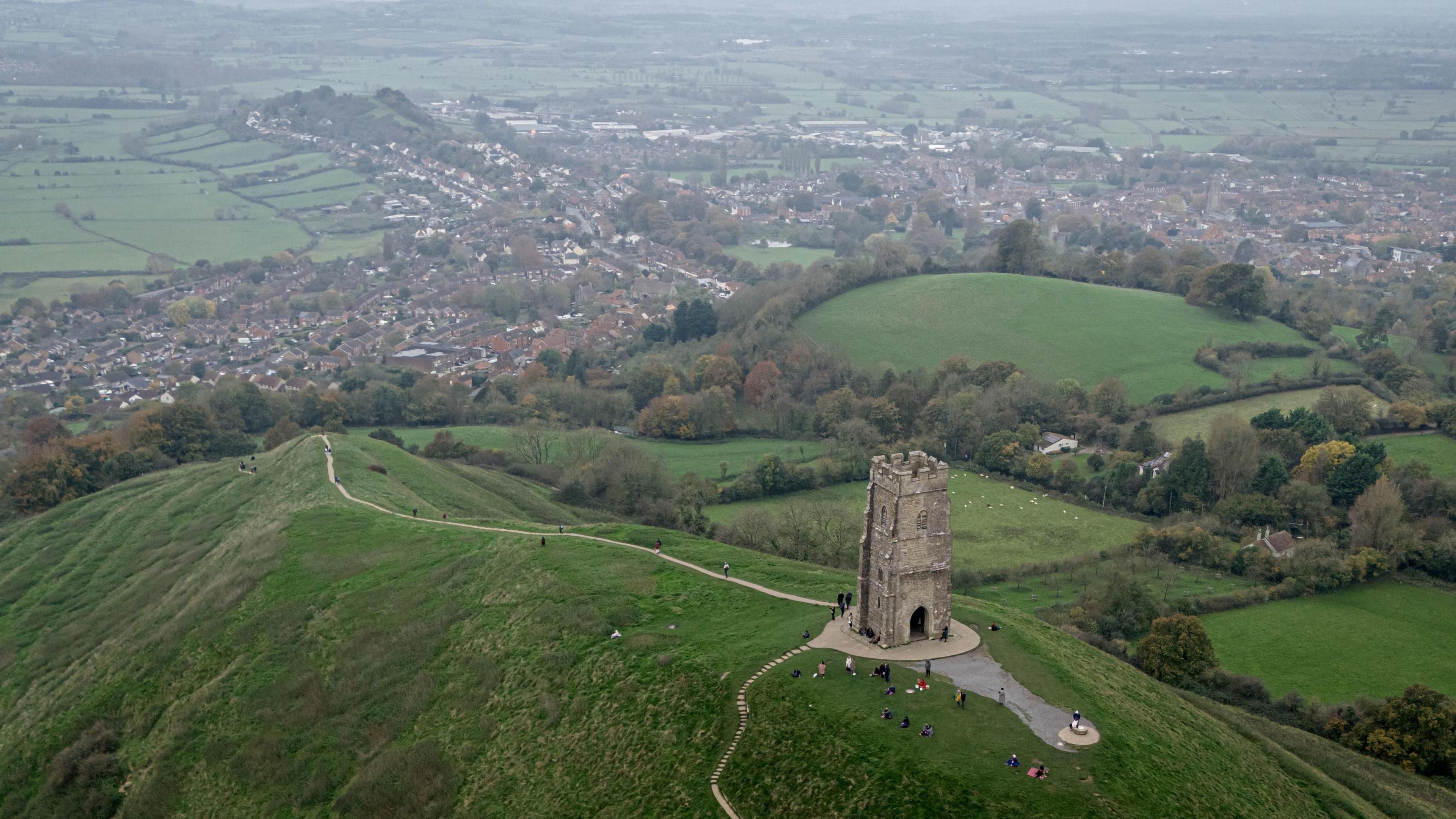 An aerial shot of Glastonbury Tor overlooking the town centre of Glastonbury in the distance. It is a slightly misty day