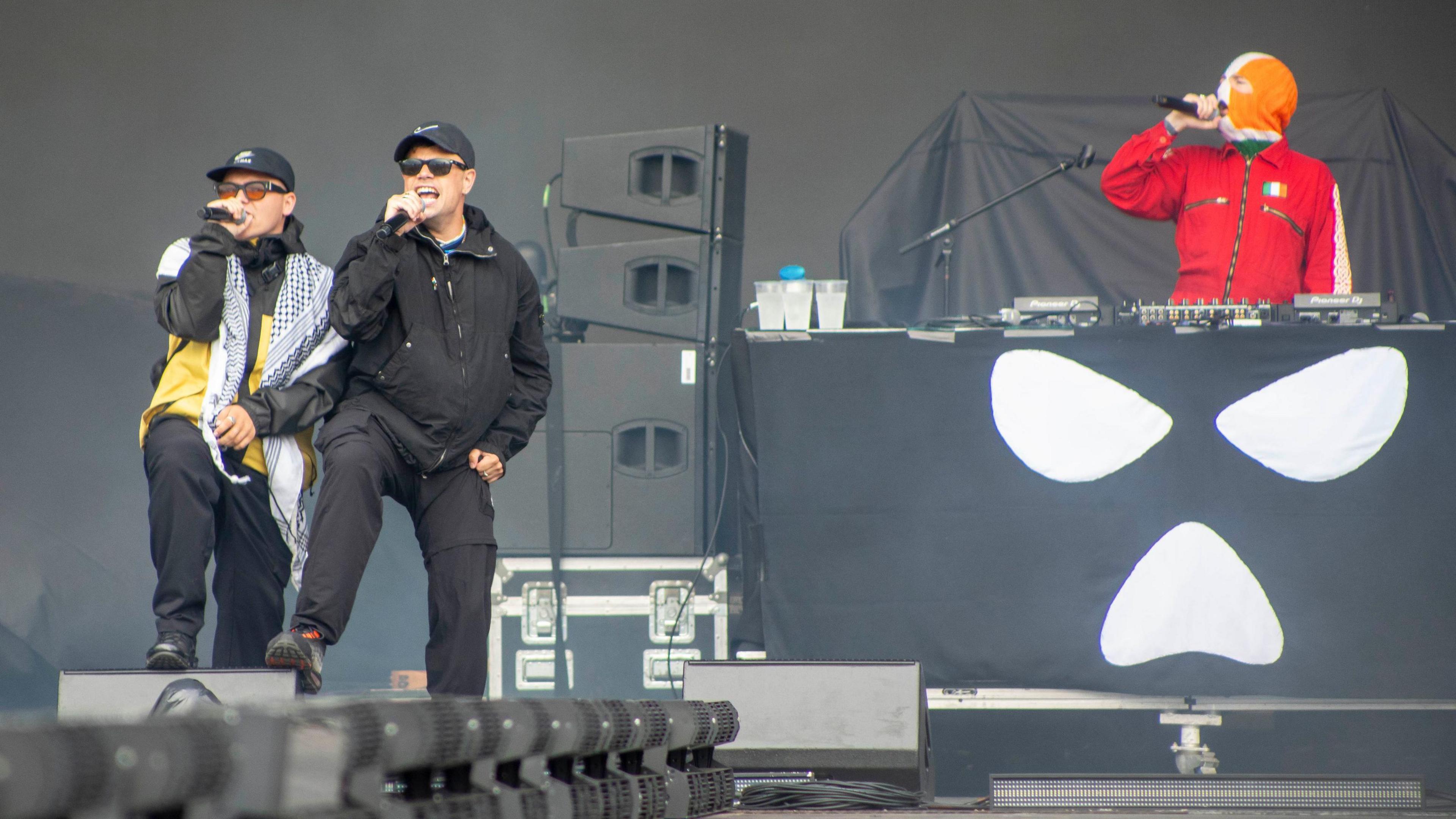 The band Kneecap perform onstage - two of the band are standing together on the left hand side, singing into microphones while wearing baseball caps and sunglasses. On the other side of the image is a DJ behind the decks, wearing a red boilersuit and balaclava in the colours of the Irish flag.
