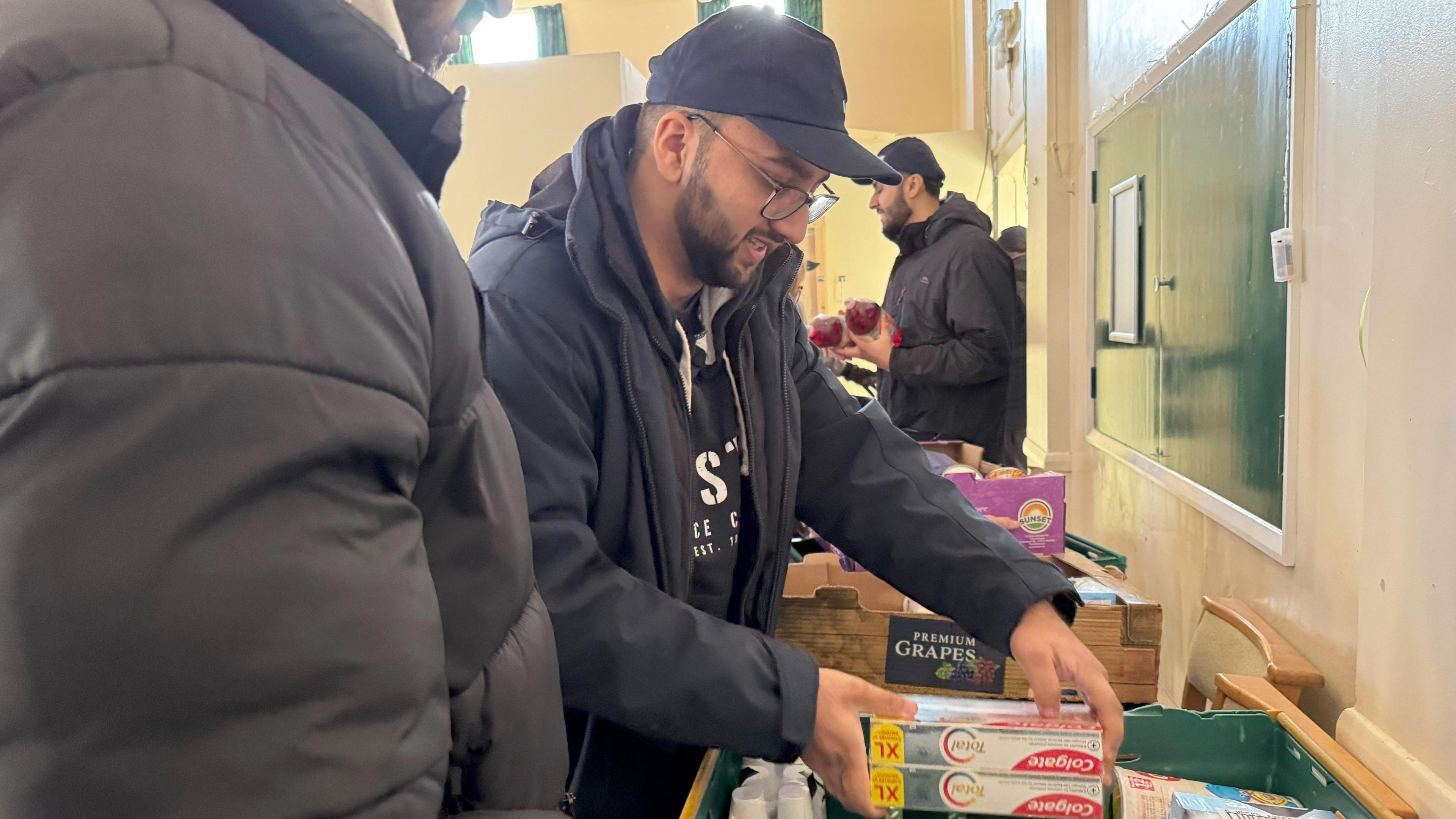 A man in a black jacket and baseball cap puts boxed toothpaste in a green crate.