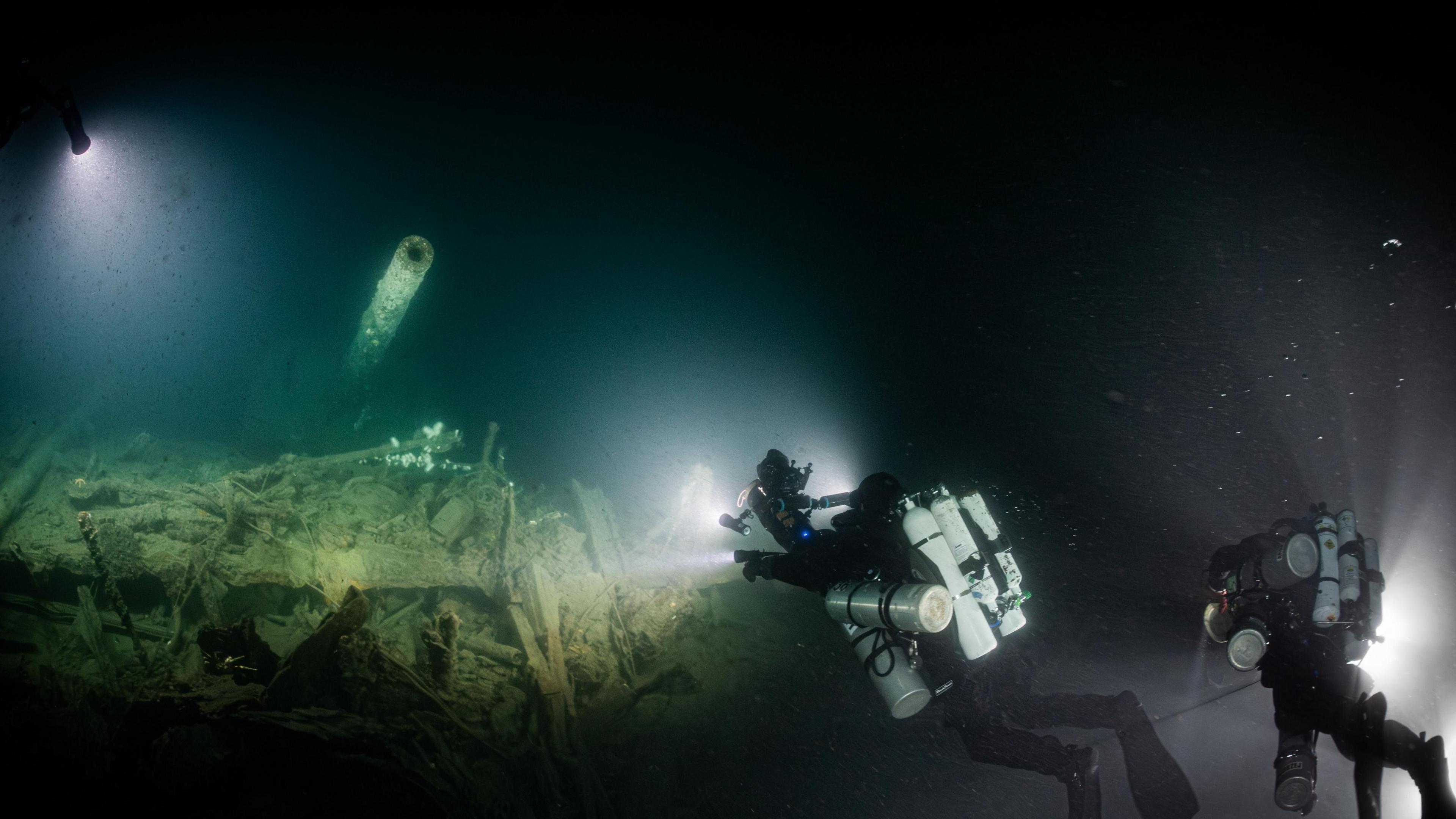 Divers shine a light on a gun turret, part of the wreckage of a Royal Navy warship.