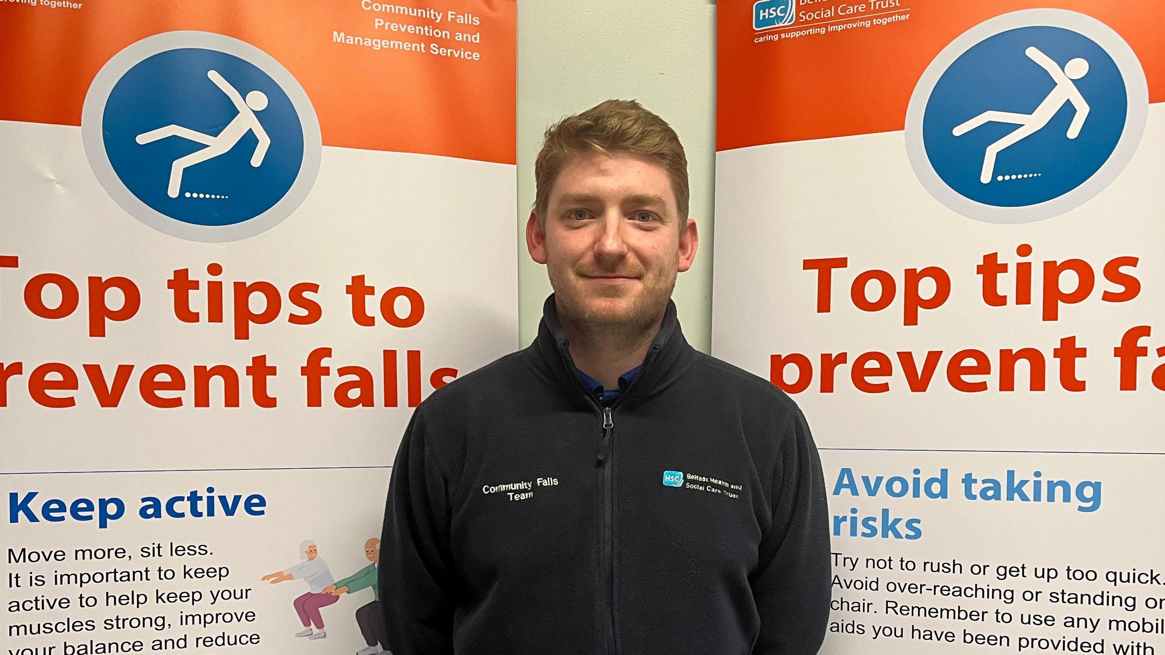 Chris McKenna smiling at the camera. He has short, fair hair and is wearing a dark Belfast Health Trust fleece jacket. There are two banners behind him displaying "top tips to prevent falls".