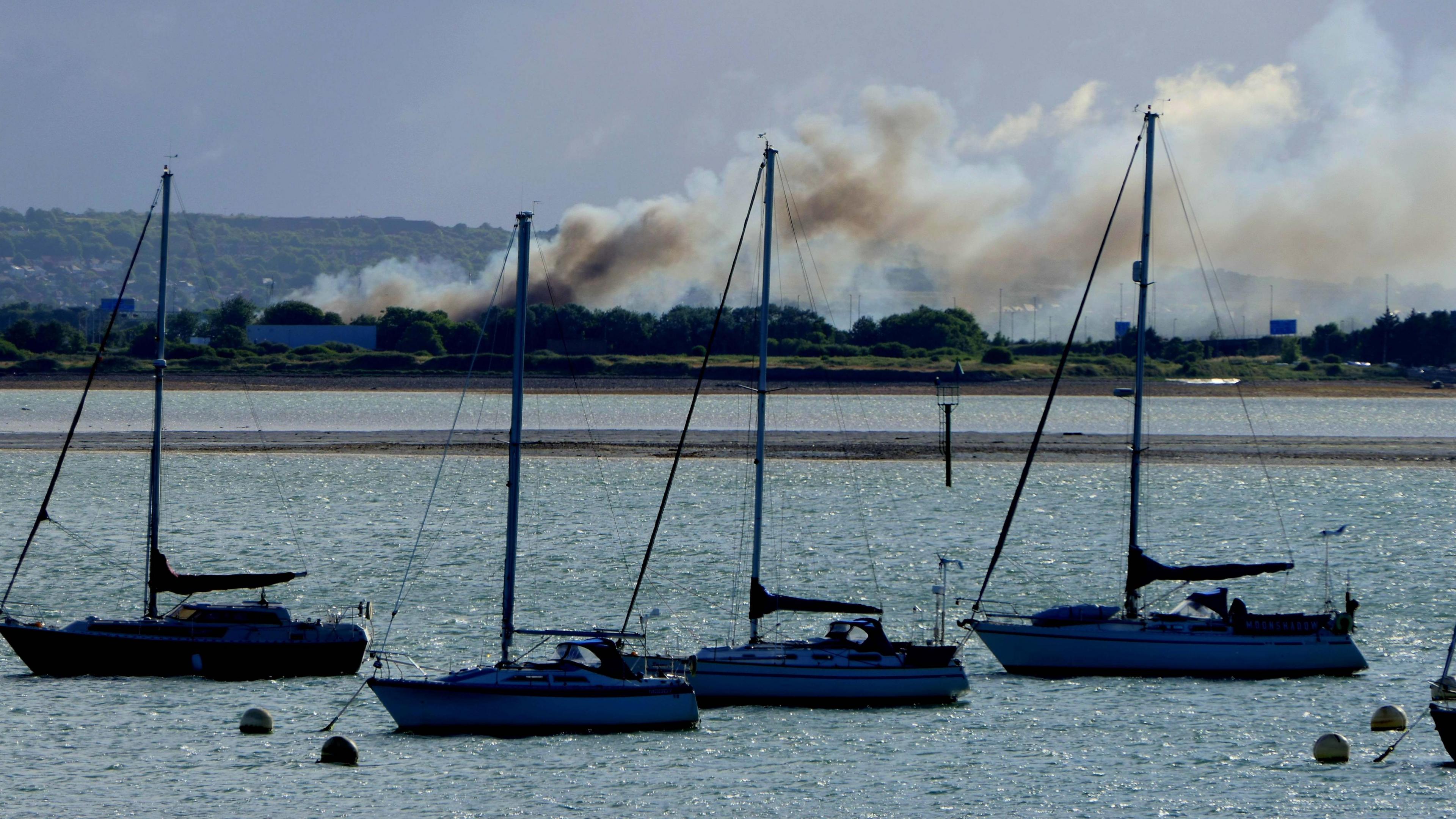 Smoke from the fire billowing across the sky, with sailboats in he foreground