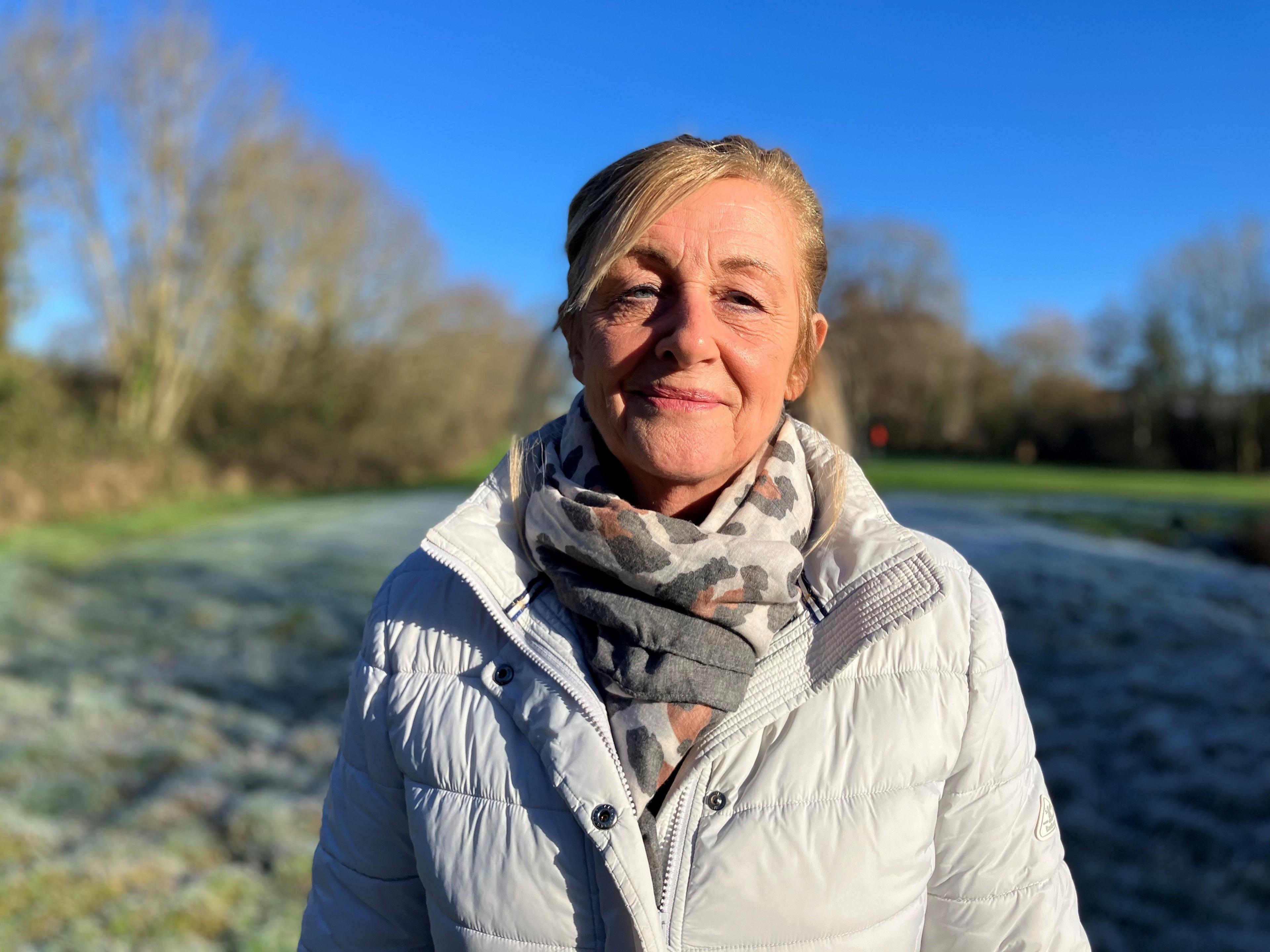 A headshot of lady in a grey coat standing in a field. She is smiling into the camera and is wearing a light-grey leopard print scarf. The sky is bright blue in the background and the outline of trees can be seen. 