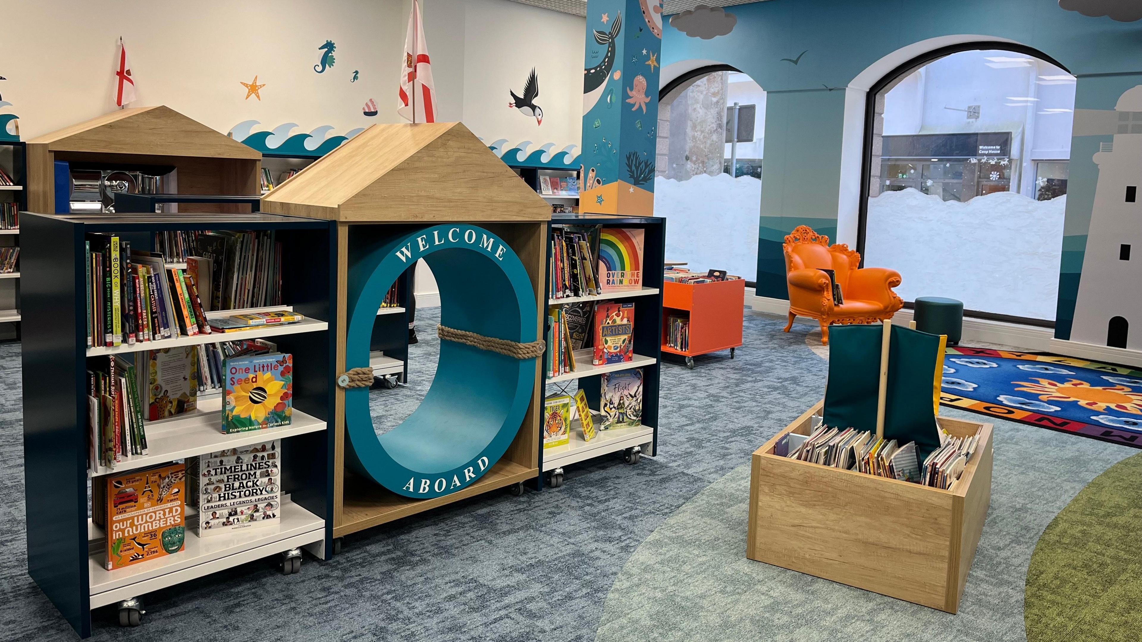 Inside Jersey's Children's Library. It is nautical themed, there is a lighthouse and sea creatures painted on the walls. An orange arm chair is in the back corner, and the bookshelves are shaped like boats. A porthole in the middle of one of the bookshelves says 'welcome aboard'.