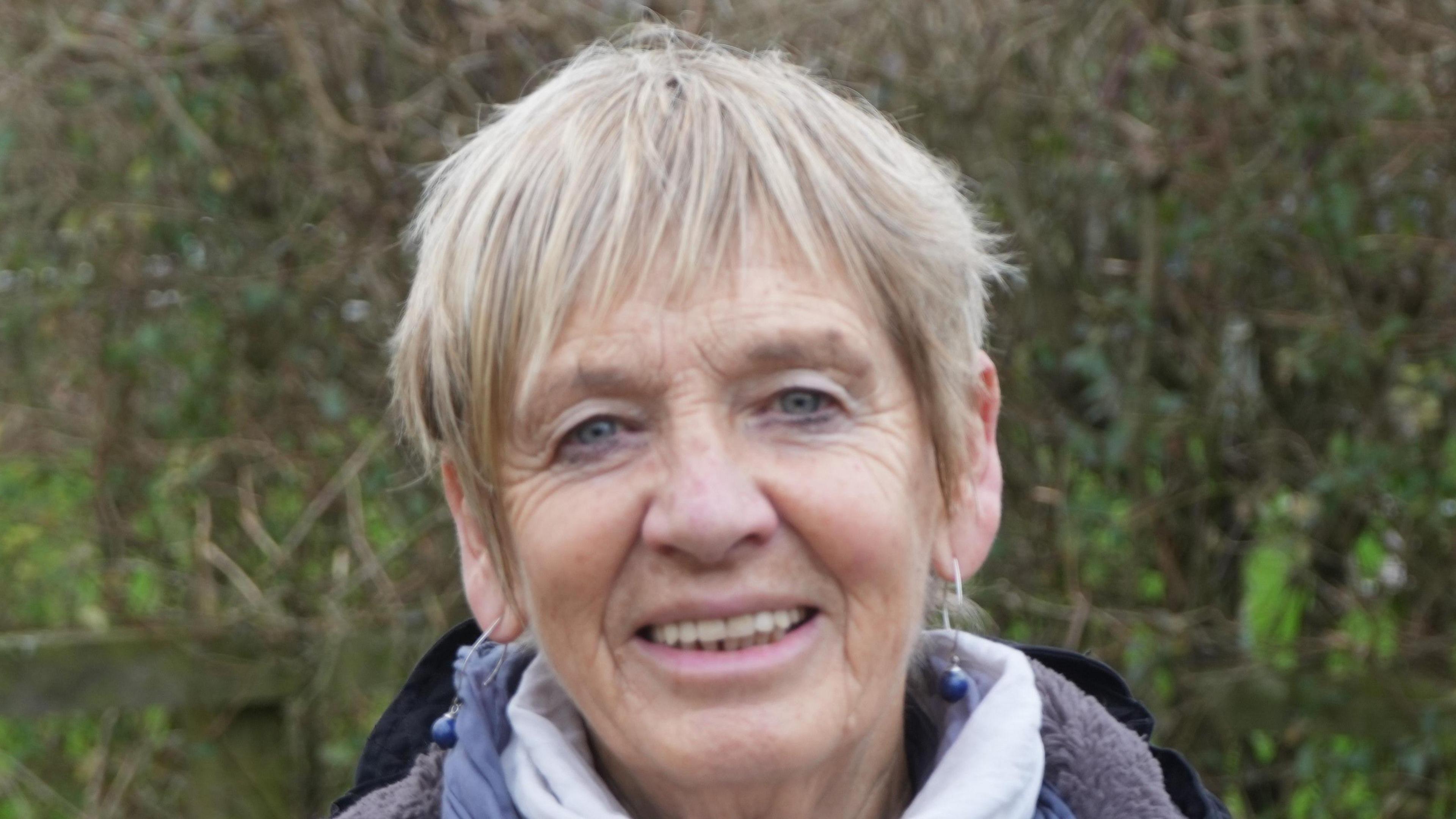 A woman with a blonde pixie hair cut smiles at the camera. Behind her is a hedge. She wears hooped earings with a blue ball on the end of each. She is also wearing a white and purple scarf around her neck. 