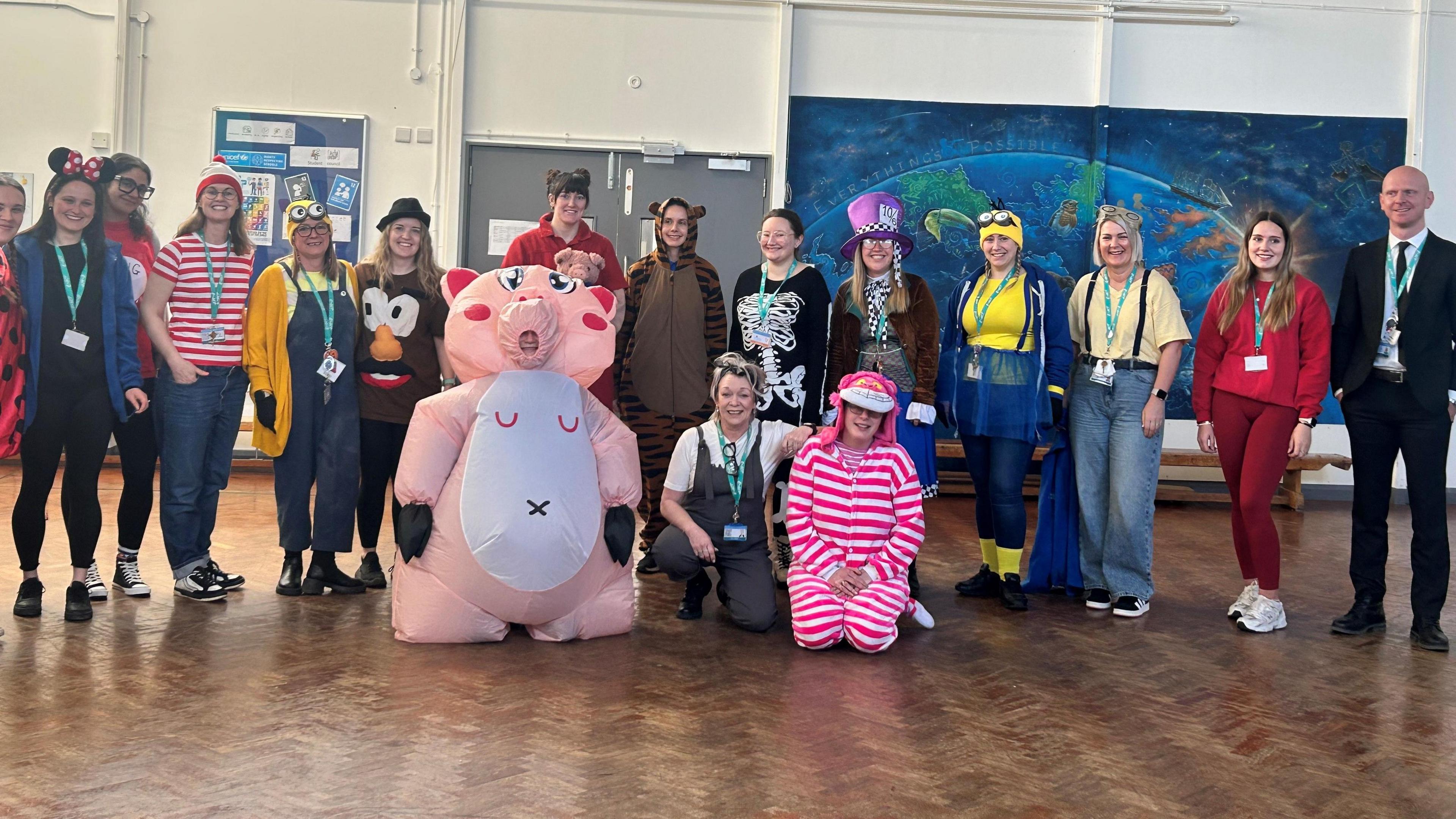 A group of teachers stand in a school gymnasium facing the camera. They are all wearing different fancy dress outfits for World Book Day