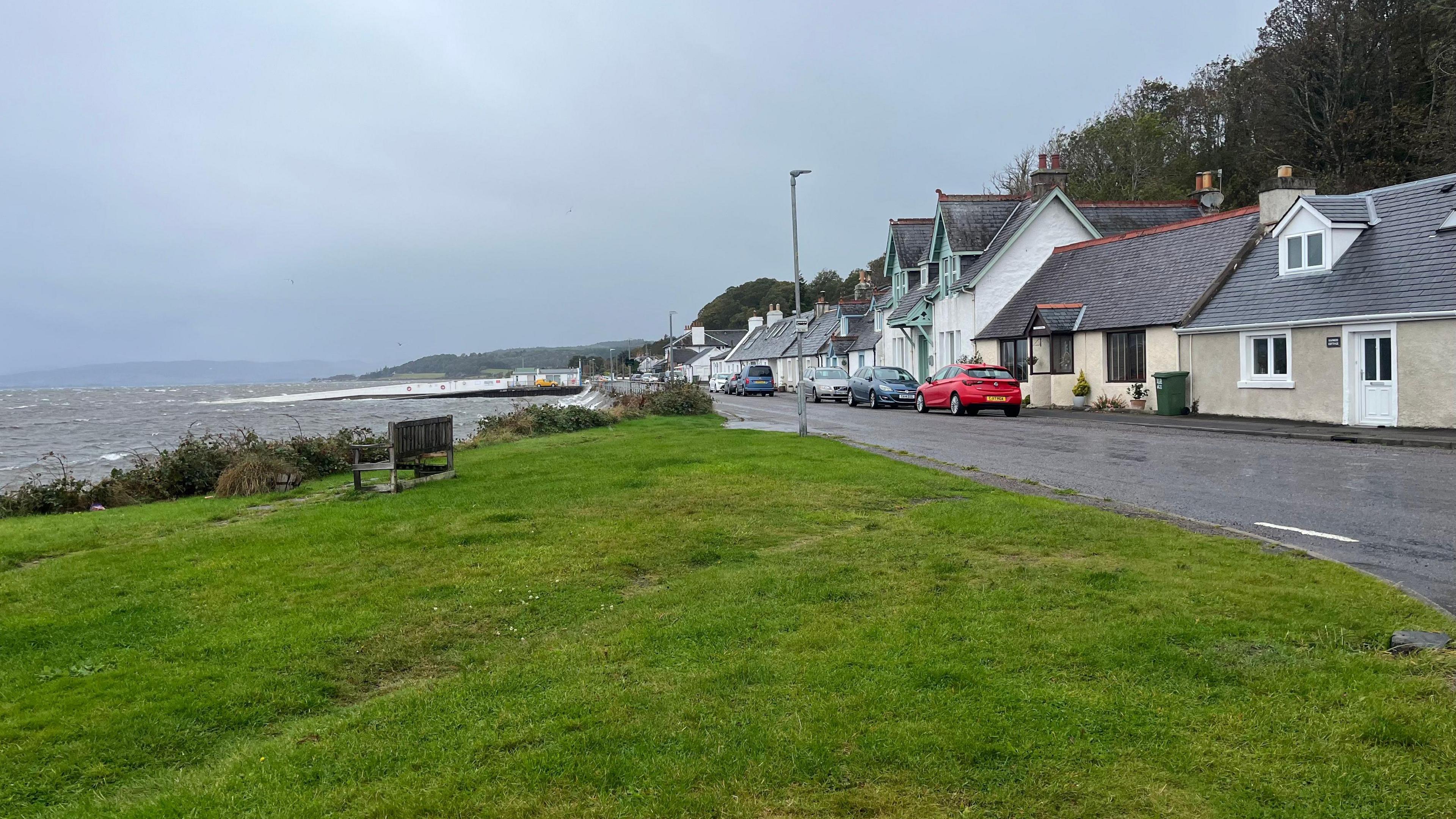 Houses line the street along the waterfront at North Kessock. It's a wet day and the Moray Firth looks grey and choppy. There is a grassy bank in the foreground, and the old ferry pier is visible in the background.