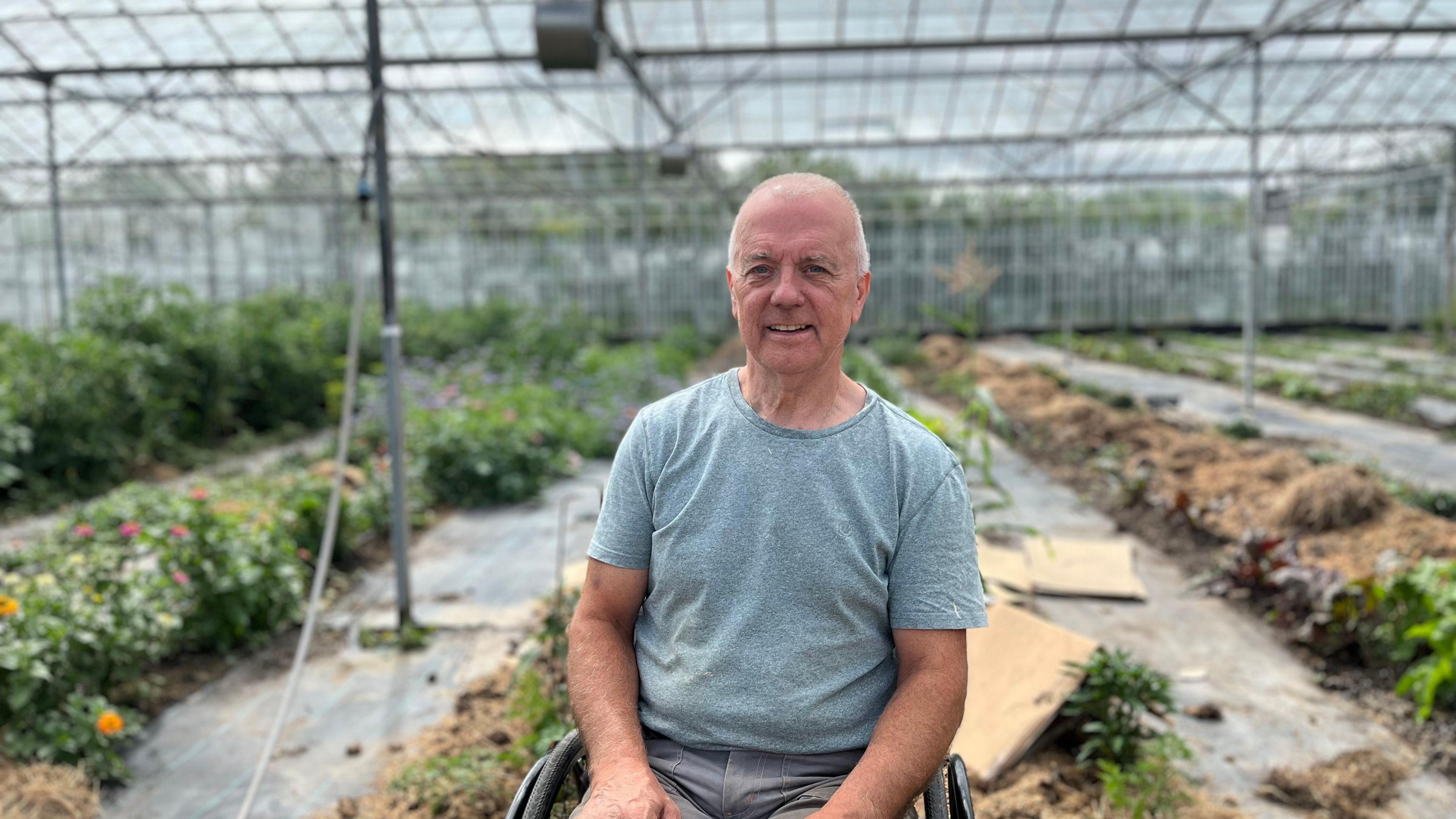 A man in a wheelchair sits in a large greenhouse full of flowers.