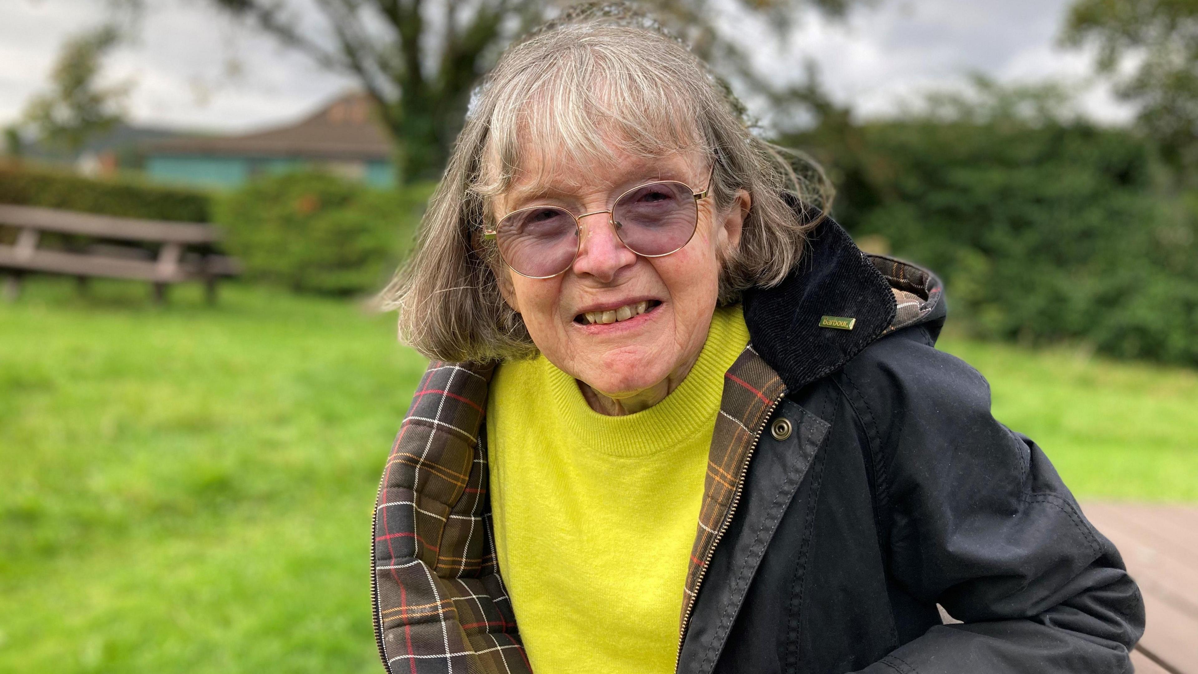 Anna Phillips looking at the camera and smiling. She is sat on a bench in the park, she is wearing a black coat with a yellow jumper underneath. She has purple tinted glasses on. 