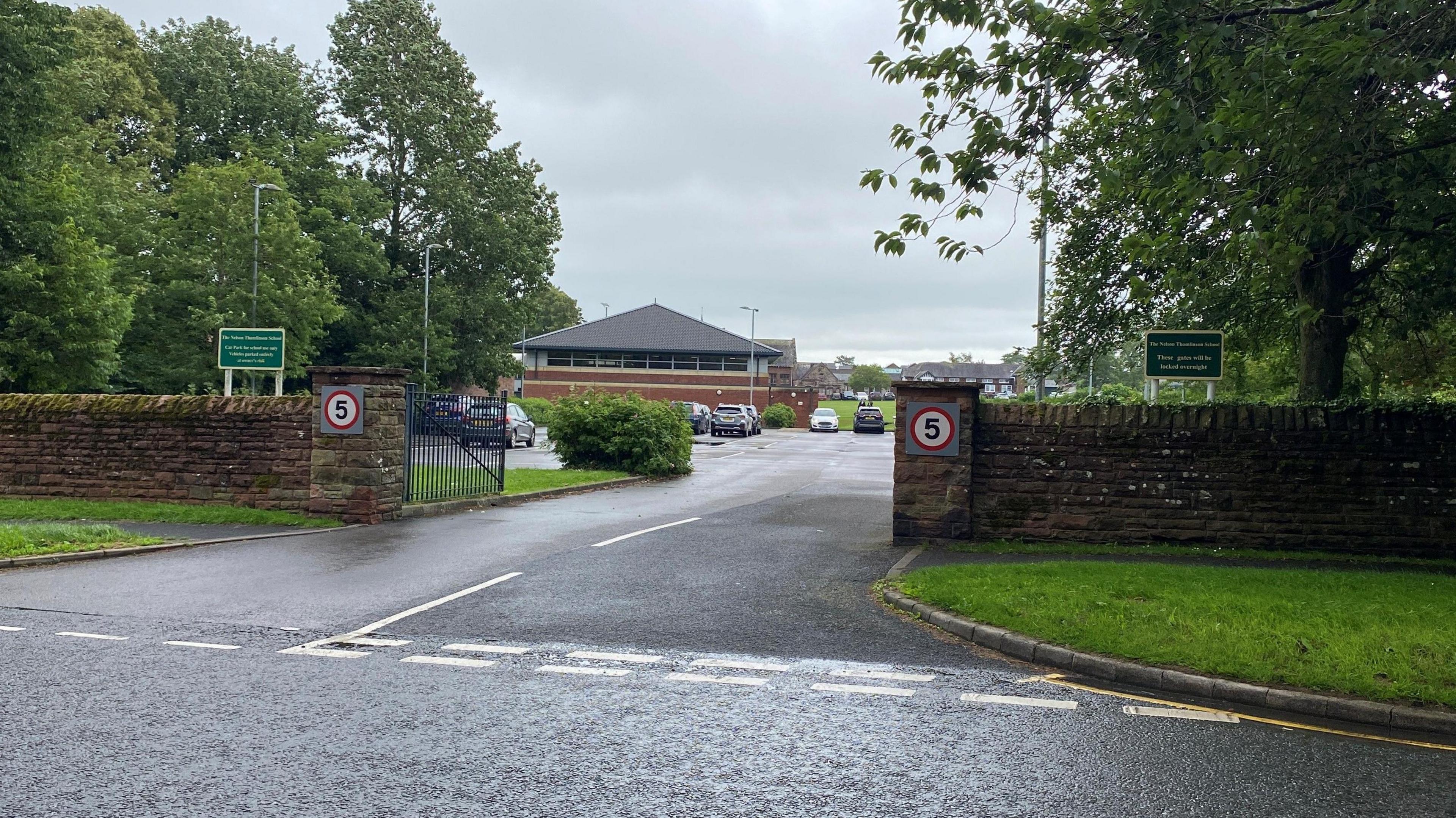 The Nelson Thomlinson secondary school taken from outside the gates shows the entrance with concrete posts on either side. The school's name is on a green sign and in the background the low school buildings which have pyramid roofs.