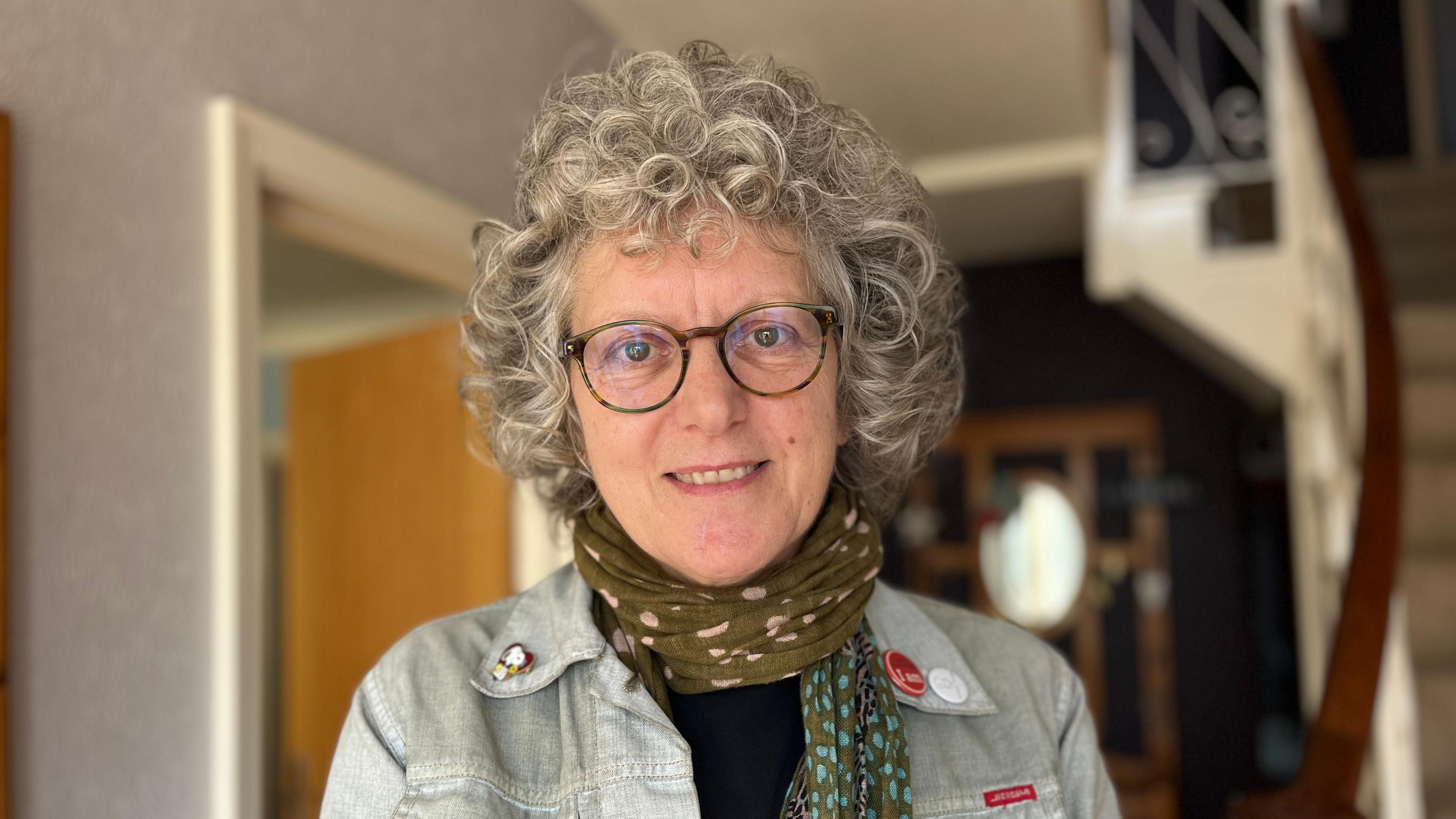 Juliette Hart is a white woman with short grey curly hair. She is wearing green glasses, a green spotted scarf and a denim jacket with badges on the lapels. She is smiling into the camera. The background is an out-of-focus house behind her.