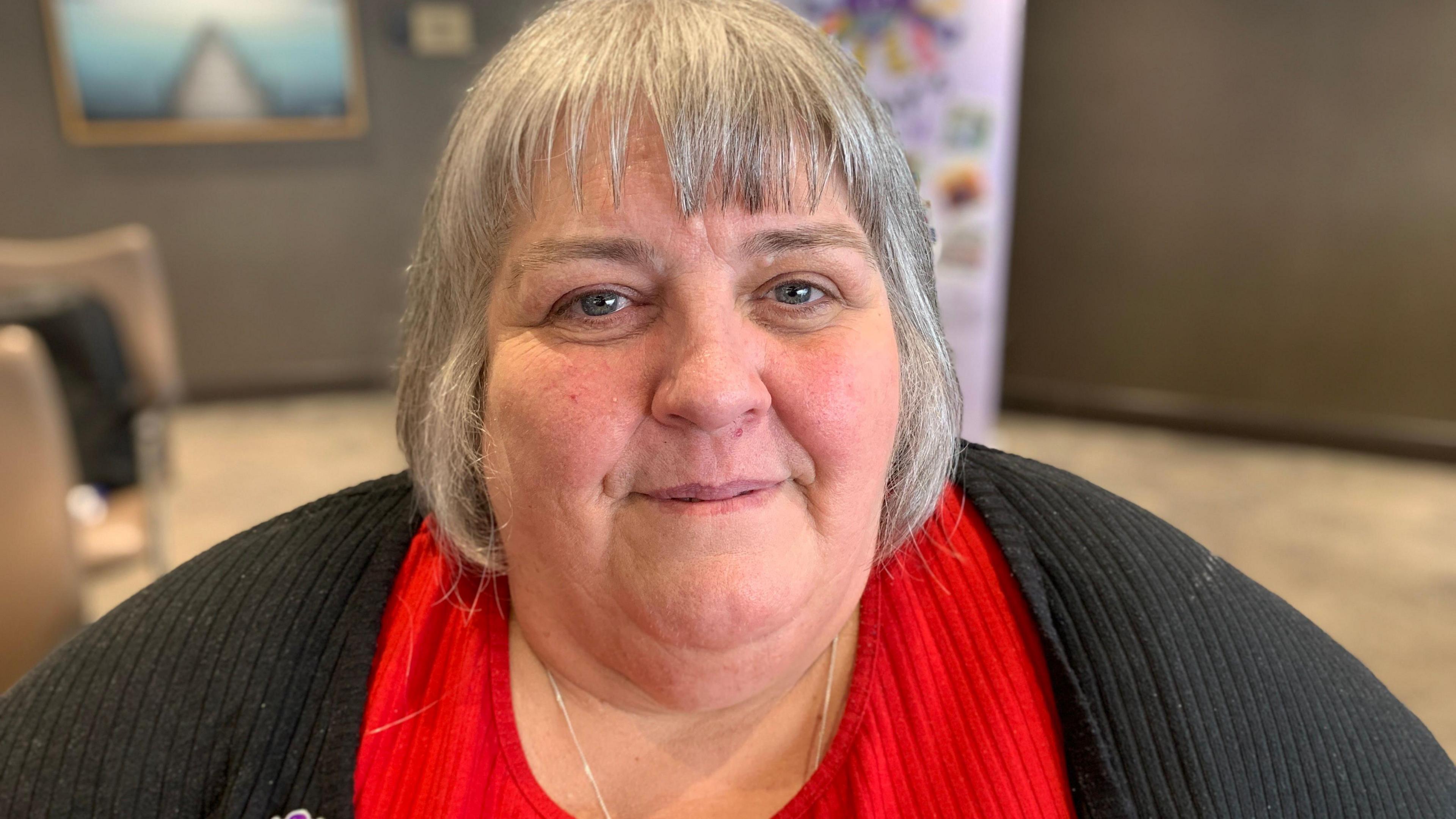 A woman with chin length grey hair is smiling at the camera. She has a black cardigan on over a red coloured top.