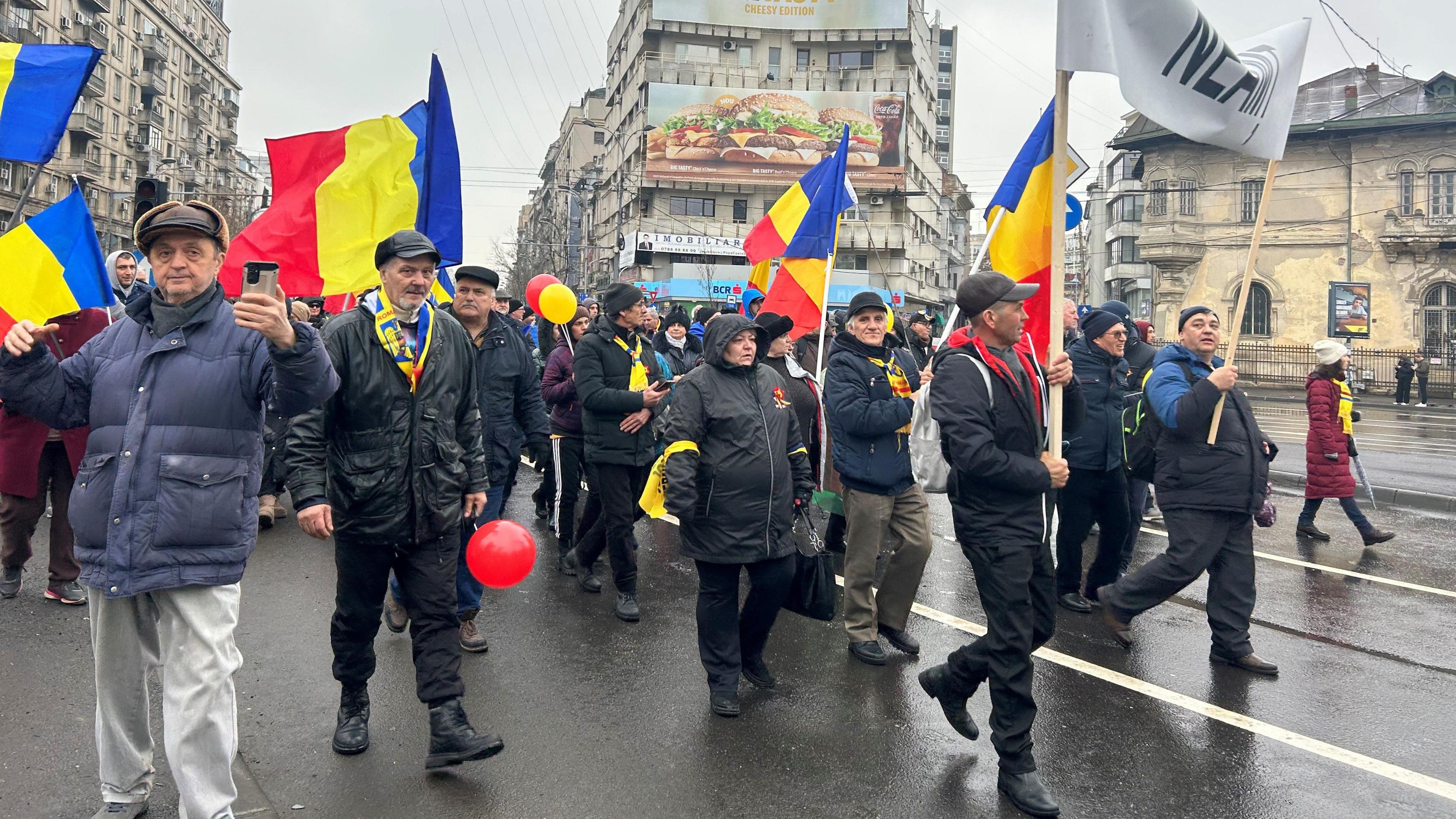 A crowd of people carrying Romanian flag march through Bucharest. Most are wearing dark clothing and hats.