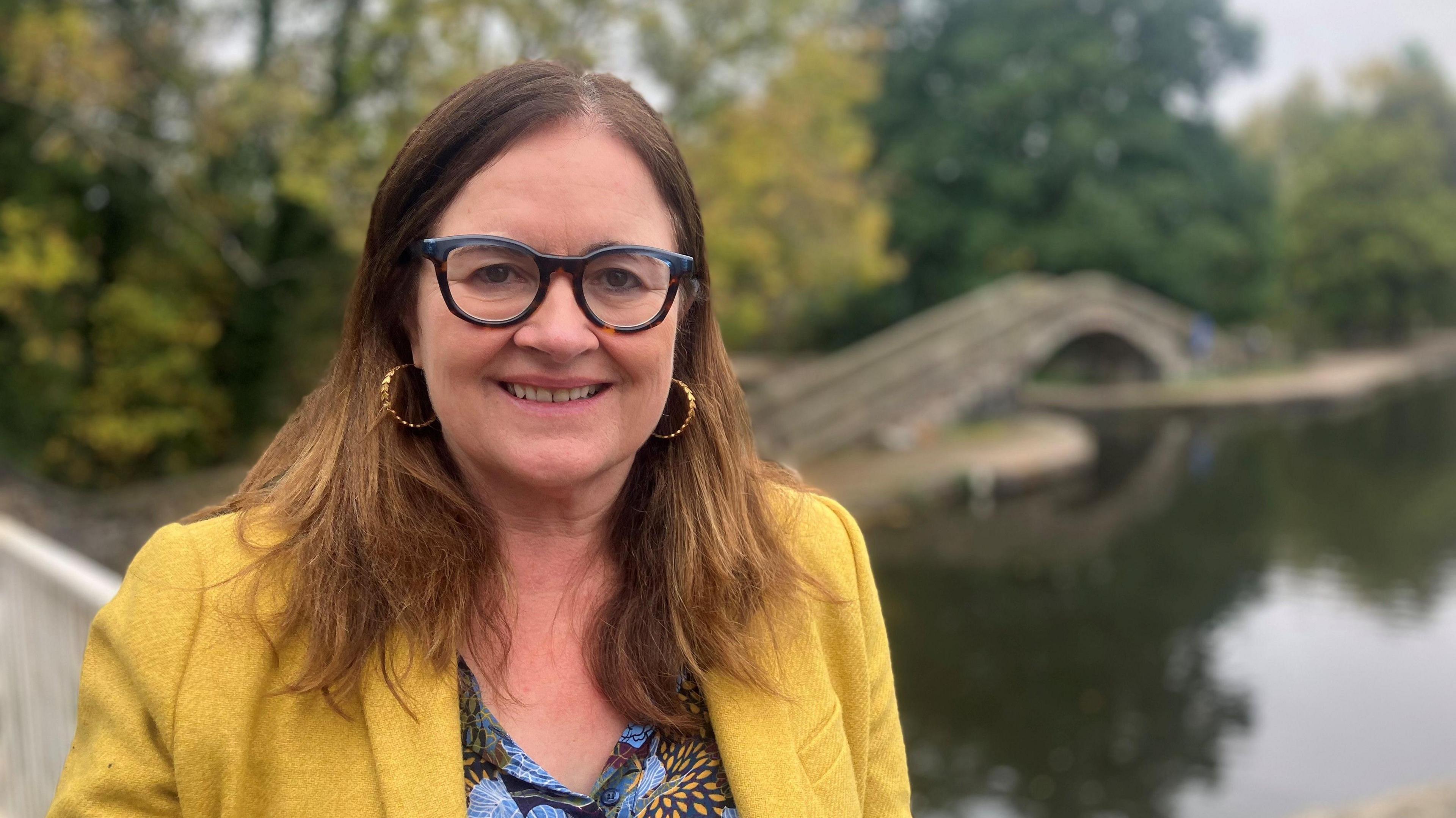 Photograph of Eilish McGuinness from the National Lottery Heritage Fund. She is pictured at the Portland Basin Museum in Ashton-under-Lyne. She has long light brown hair and wears glasses. She is wearing a yellow jacket over a blue and yellow patterned top.