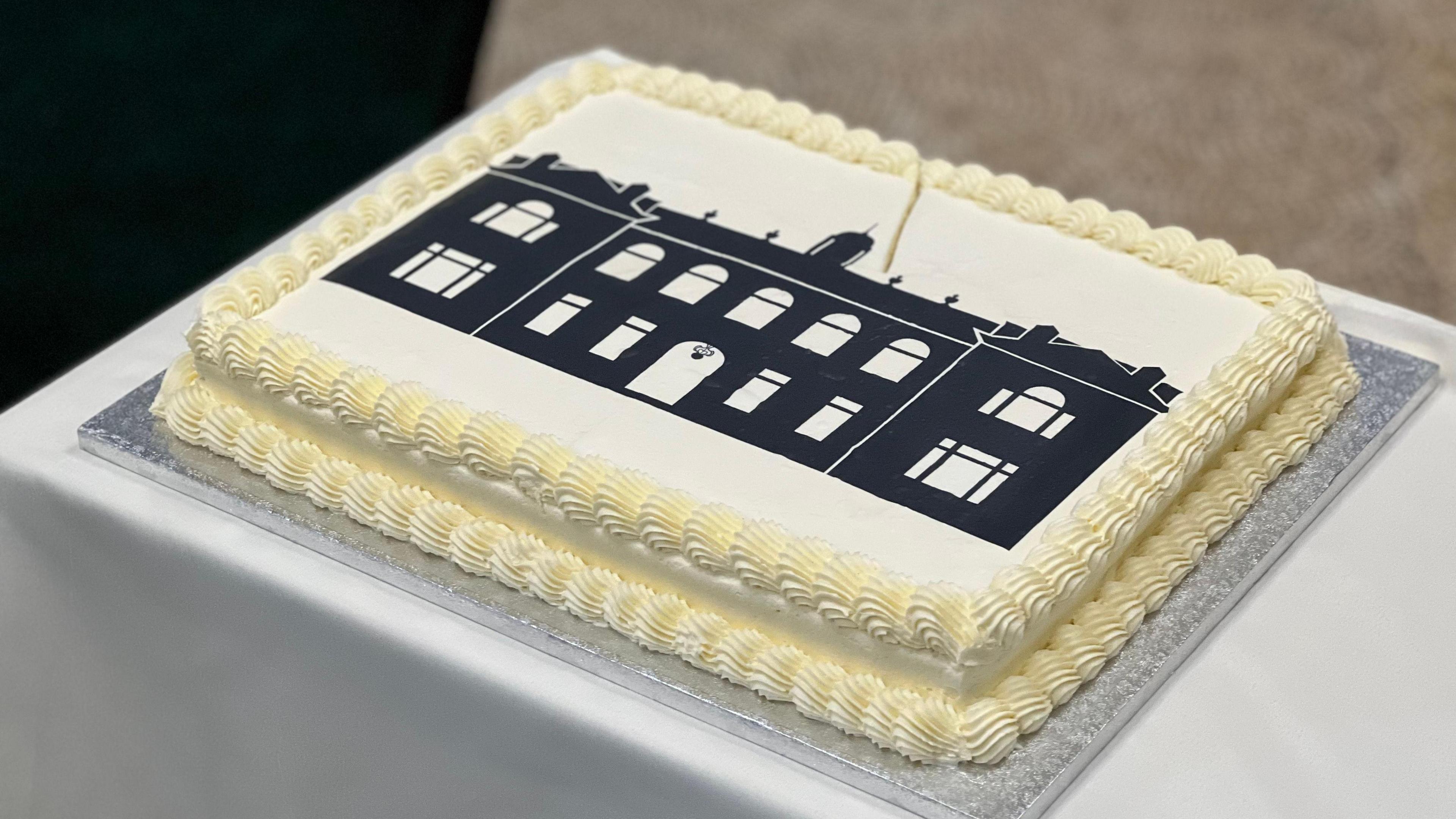 A one-layered rectangular cake covered in white icing with piping round the edges. On top, an silhouette of the shape of the building.