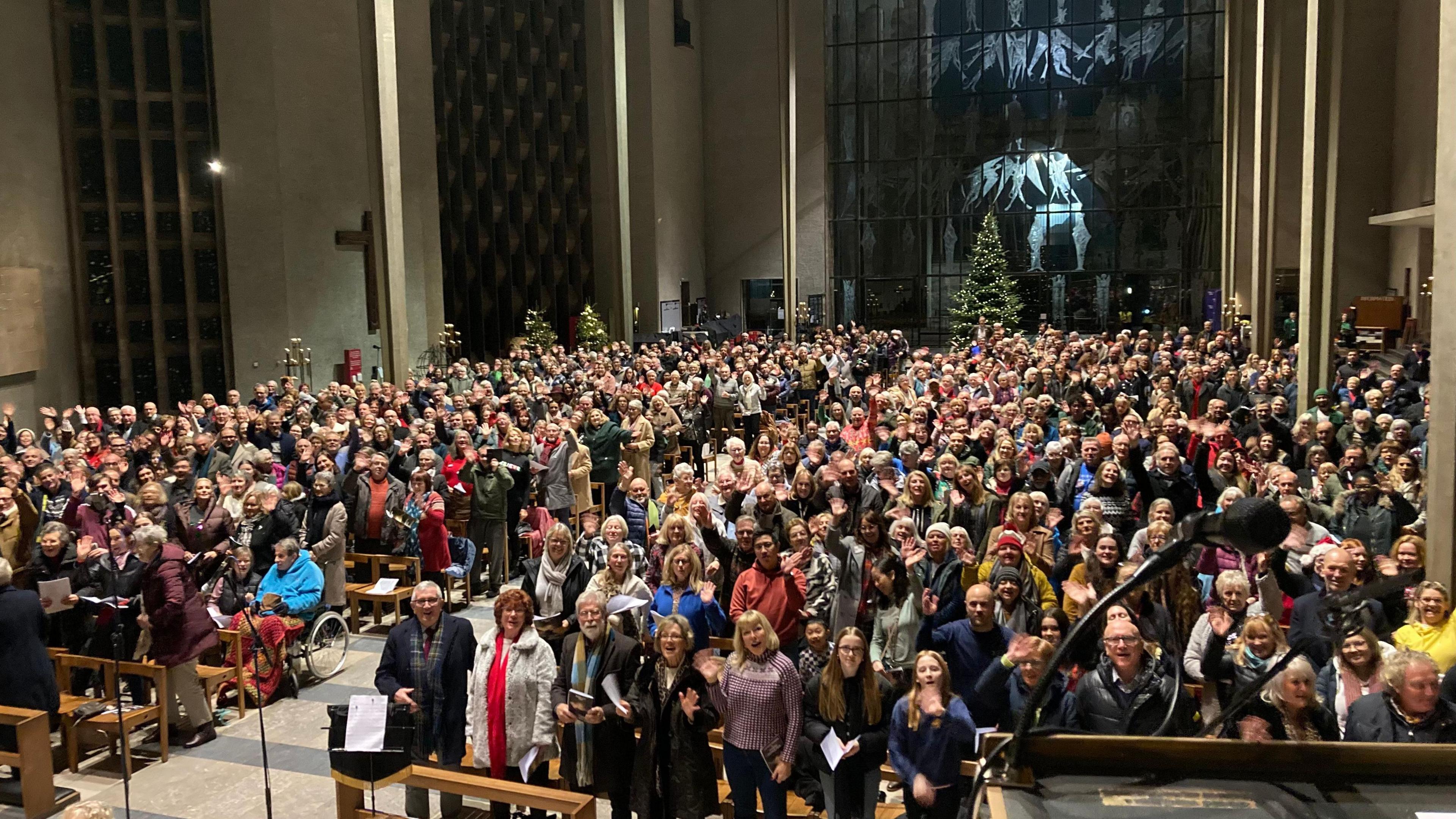 A large crowd inside the vaunted central nave of Coventry Cathedral with them all wearing jumpers and coats. There are glass windows at the back, engraved with images of angels.