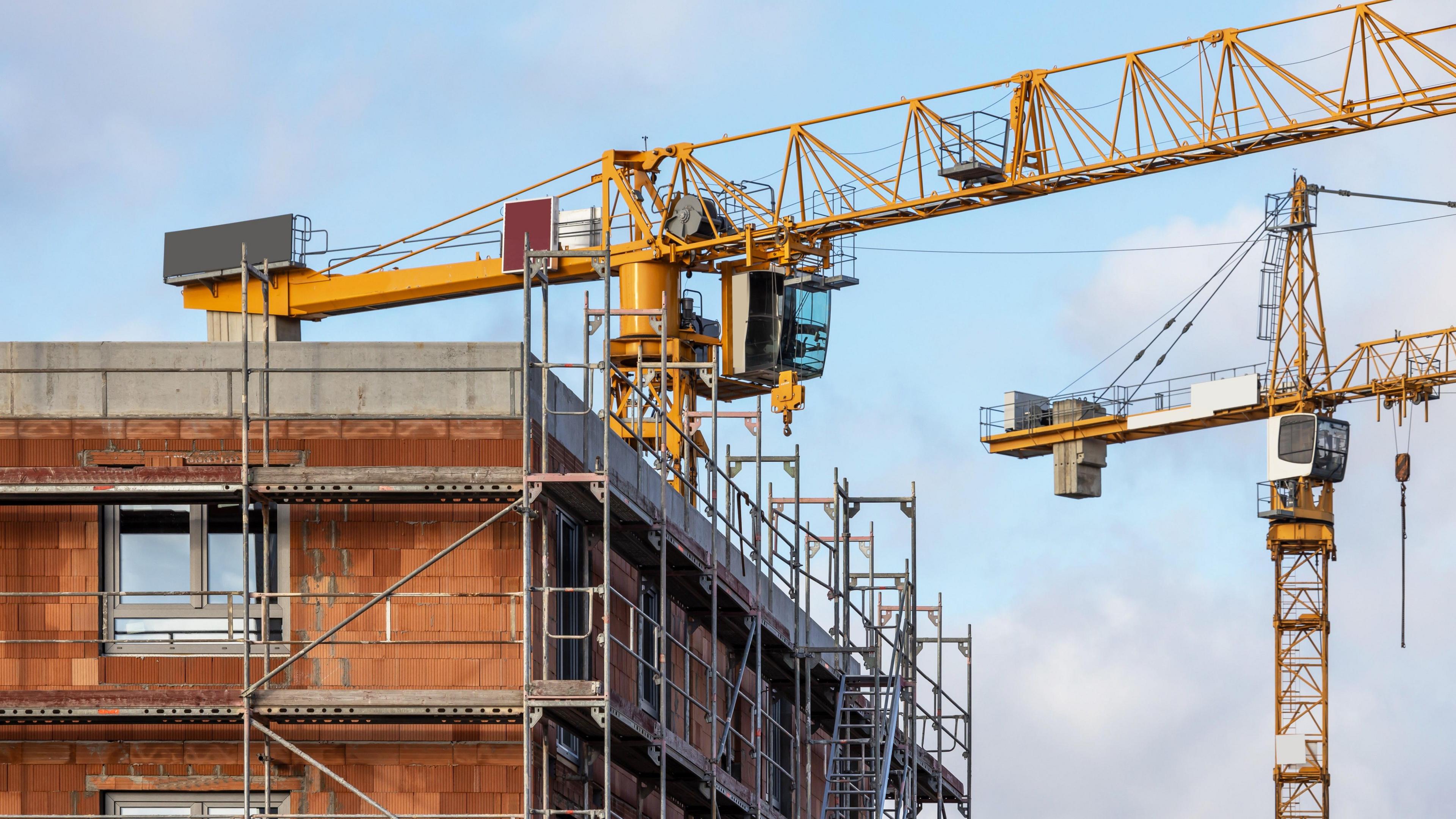 Two cranes set against a blue sky, above a partially build block, surrounded by scaffolding