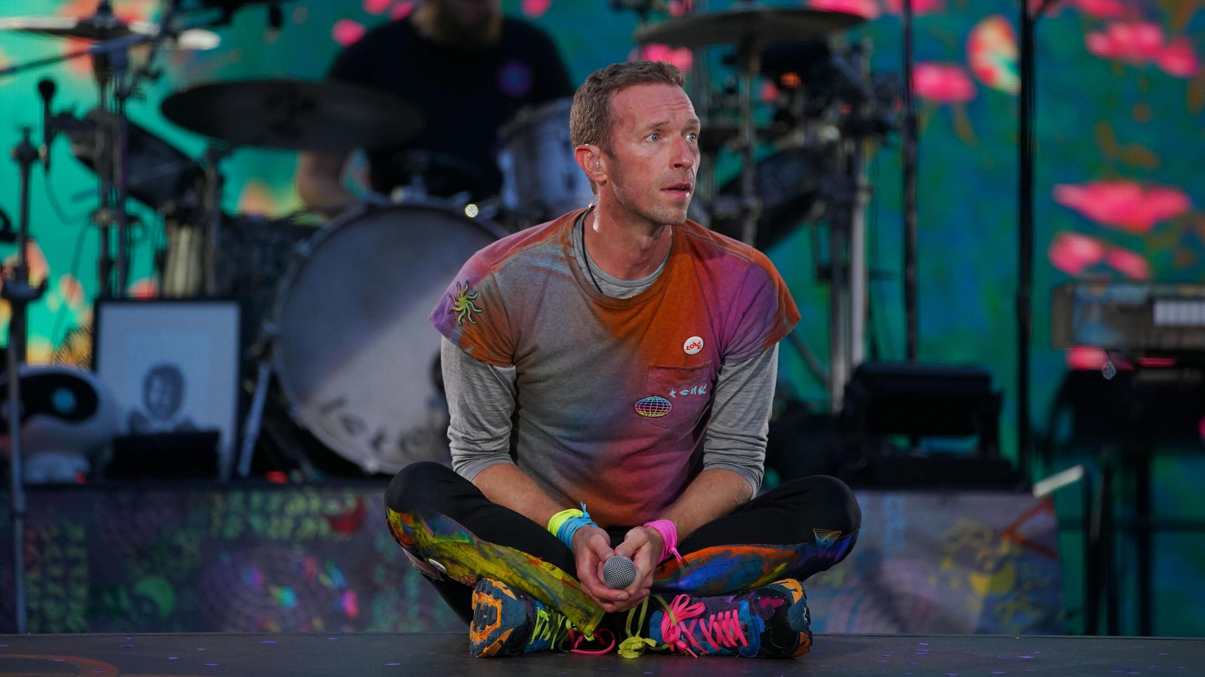 Chris Martin of Coldplay, he sits on the stage holding a microphone. He is wearing an outfit covered in ultra-violet paint splatters.