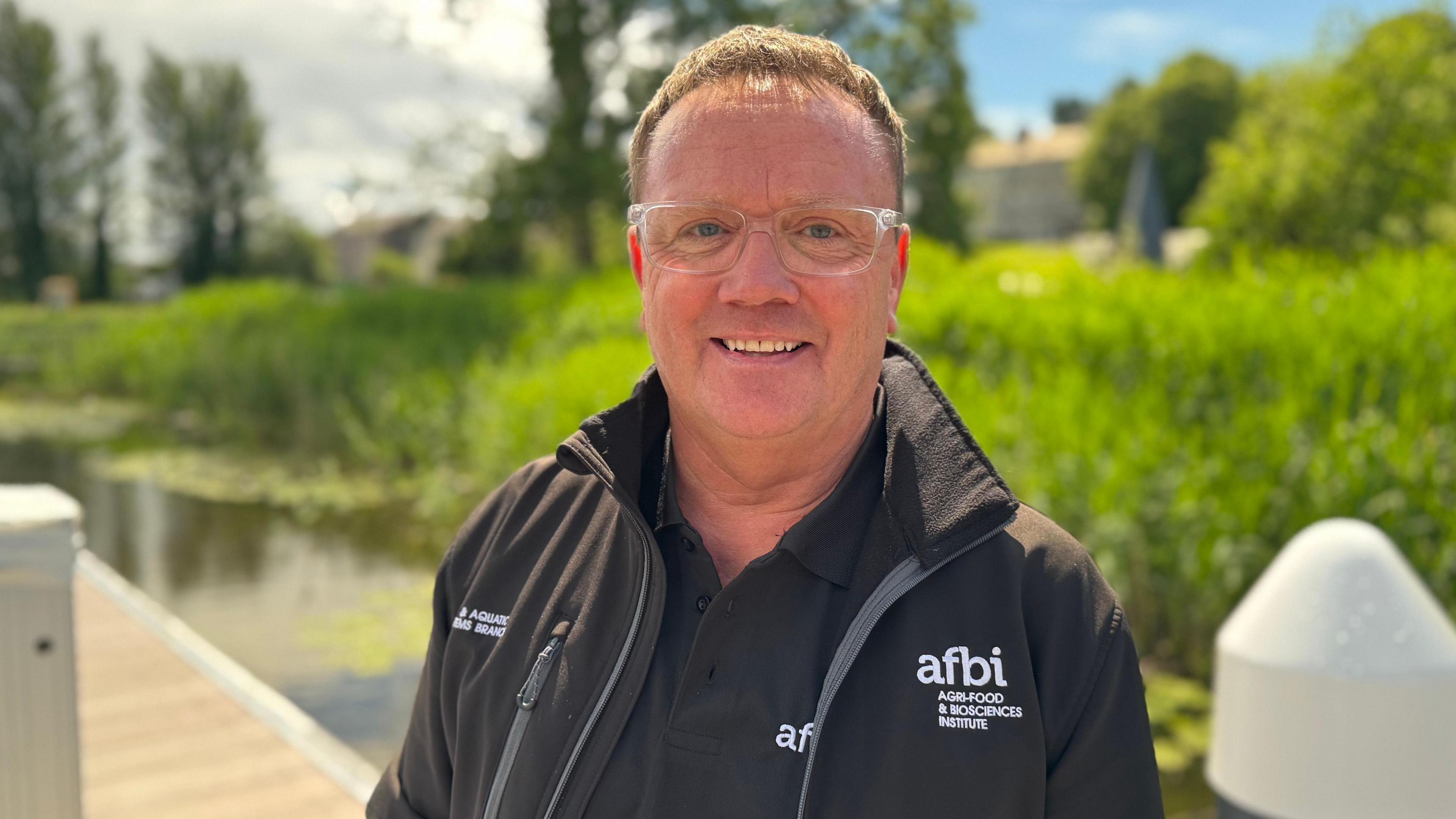 A man is wearing glasses and smiling. He's outside. A pond is behind him with foliage. He has a top and jacket that says AFBI Agri-food & biosciences institute. 