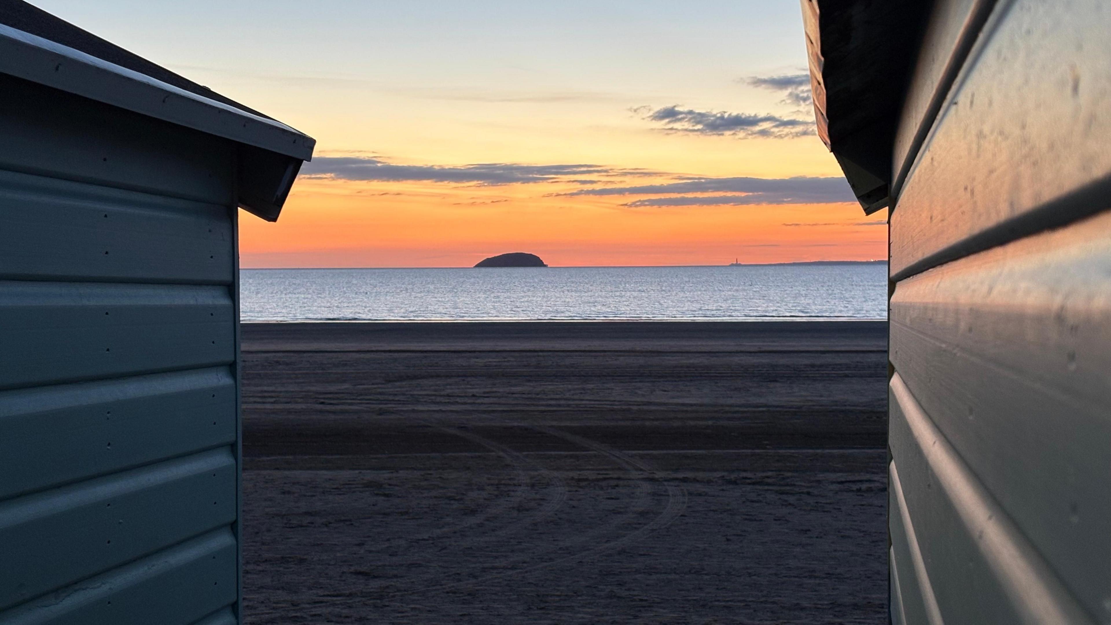 Steepholm Island, a small hill-shaped piece of land, pictured between two beach huts in Weston-super-Mare. The sand  stretches out in front of the huts, with tyre marks visible in it. The tide is in with the island visible on the horizon under a bright orange sky.