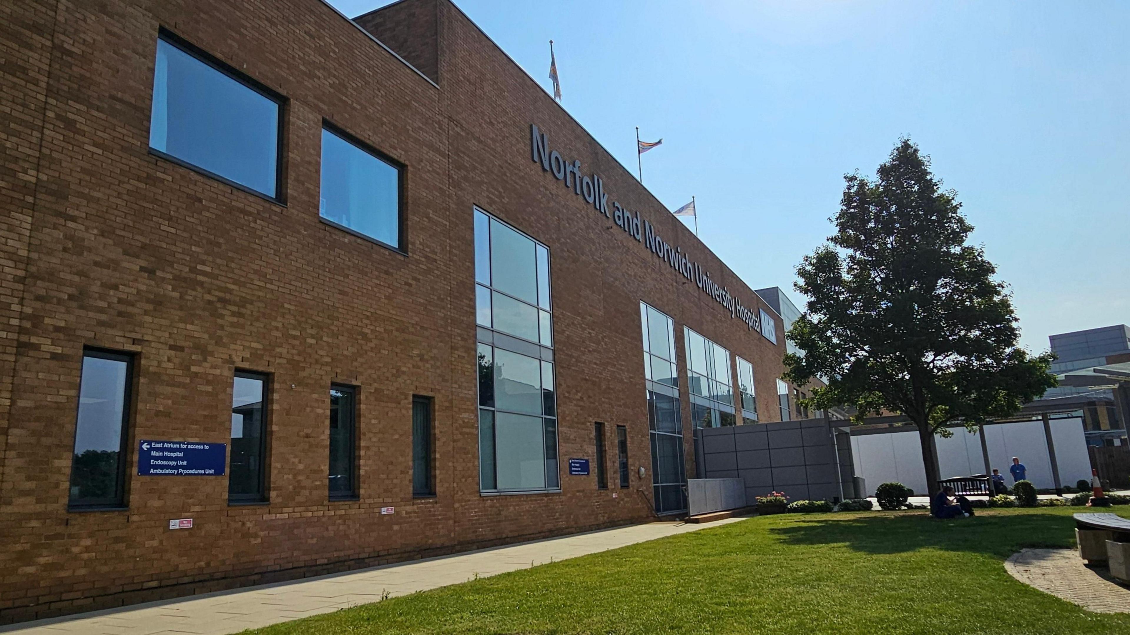 An external view of Norfolk and Norwich University Hospital, with its name high on the wall. A tree stands outside the hospital and the sky is bright blue