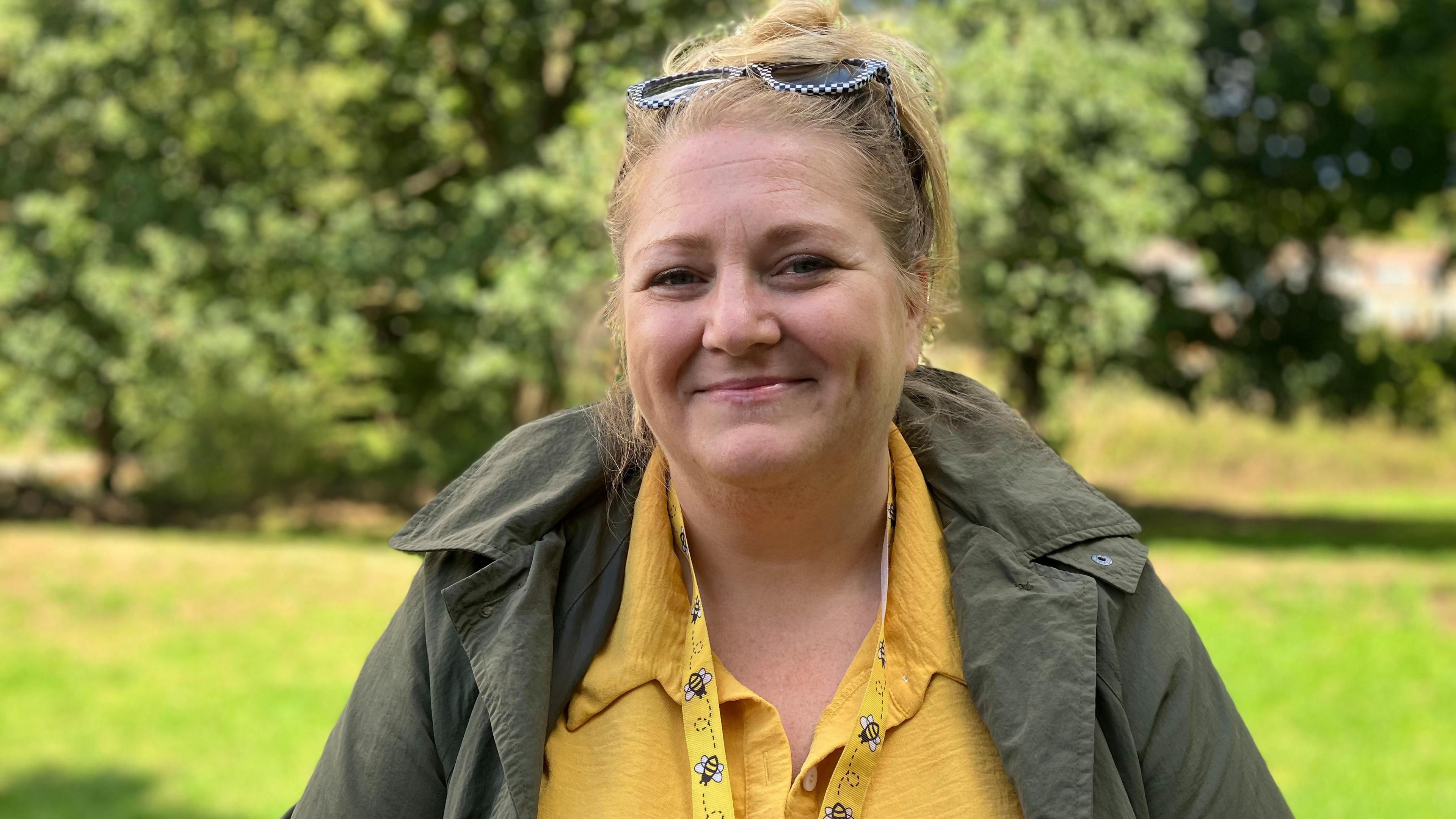 A woman smiling at the camera sitting in a park on a sunny day. She is wearing a yellow lanyard with bumblebees on it.