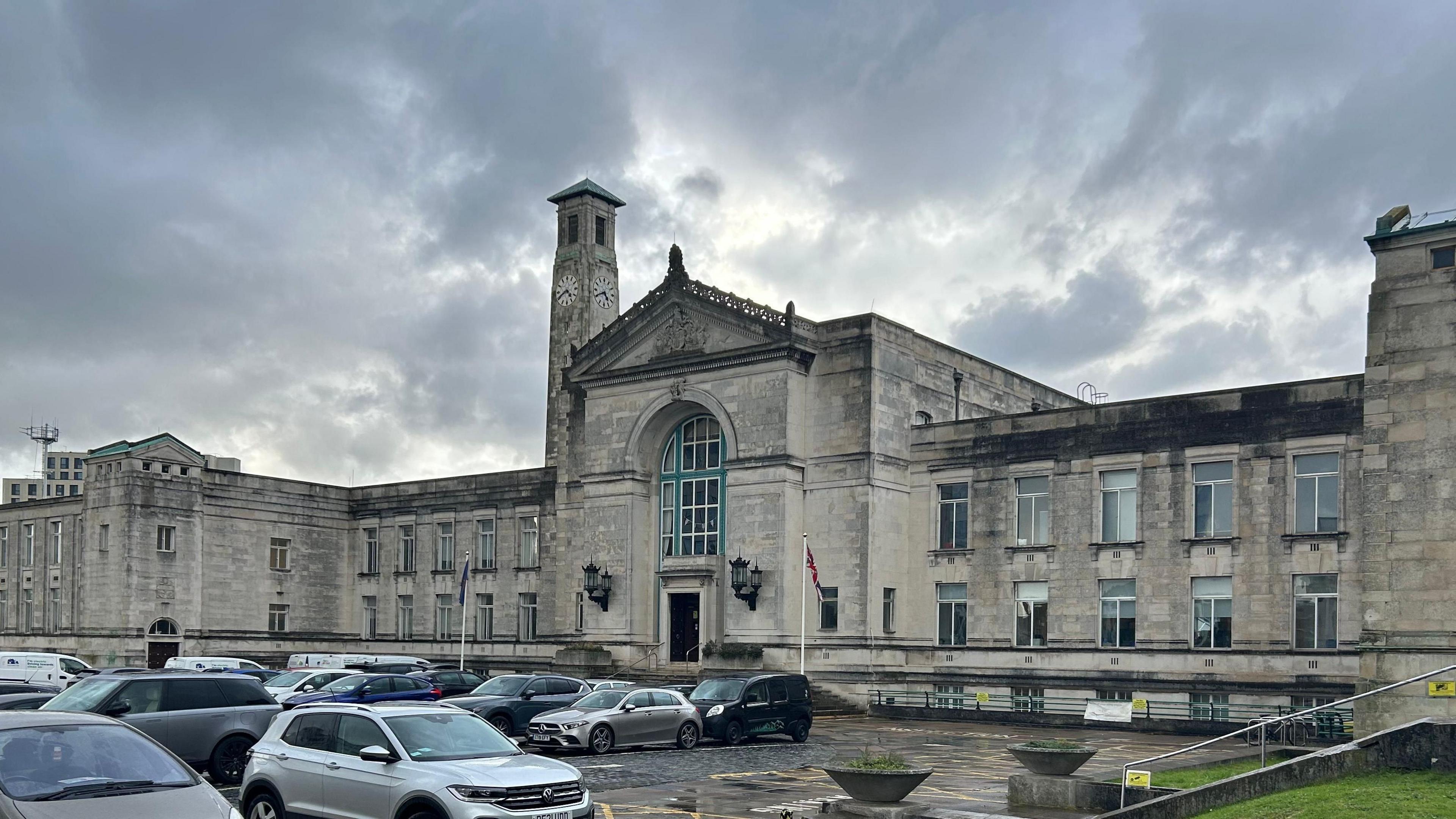 Dark clouds hover above Southampton City Council's white stone headquarters. An ornate entrance in a car park includes a tall window above the main door. The landmark civic centre clock tower is set into the sprawling building complex.