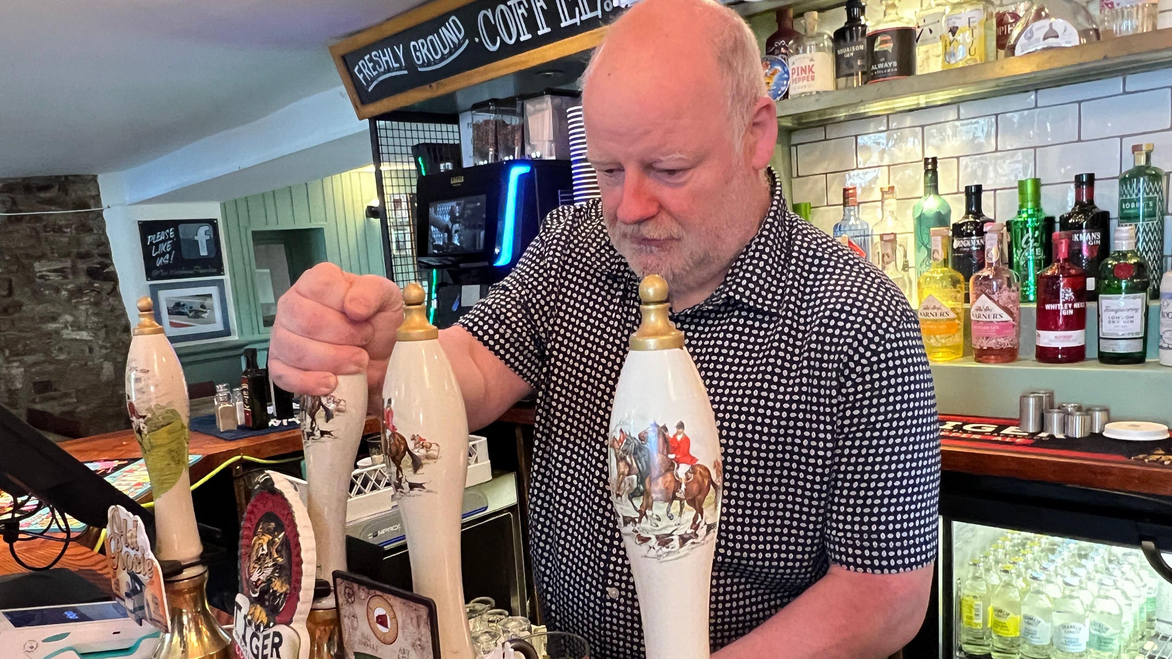 Malcolm Gray with short grey hair and beard pulling a pint in his pub