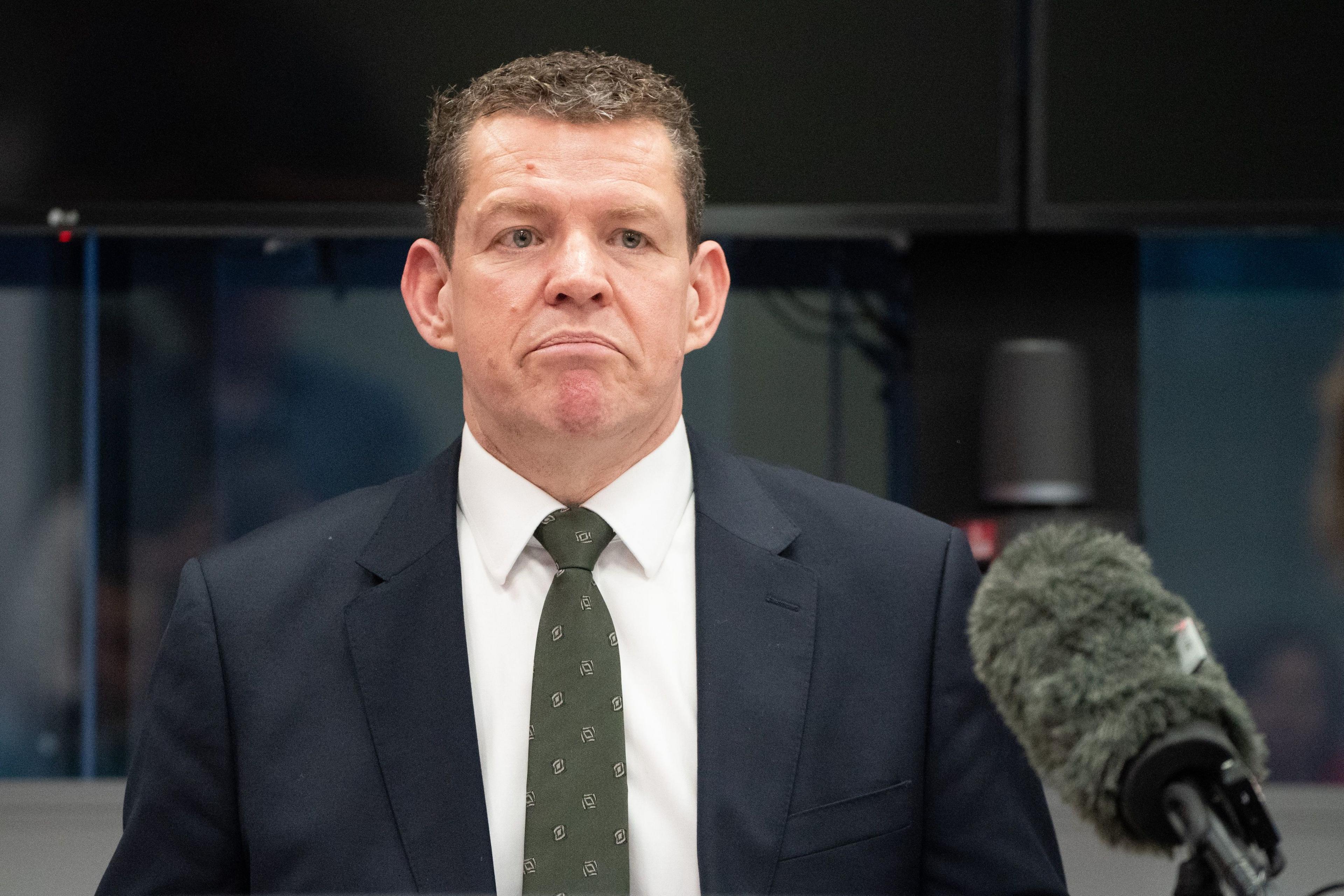 A man with short dark hair, wearing a dark suit, white shirt and green tie