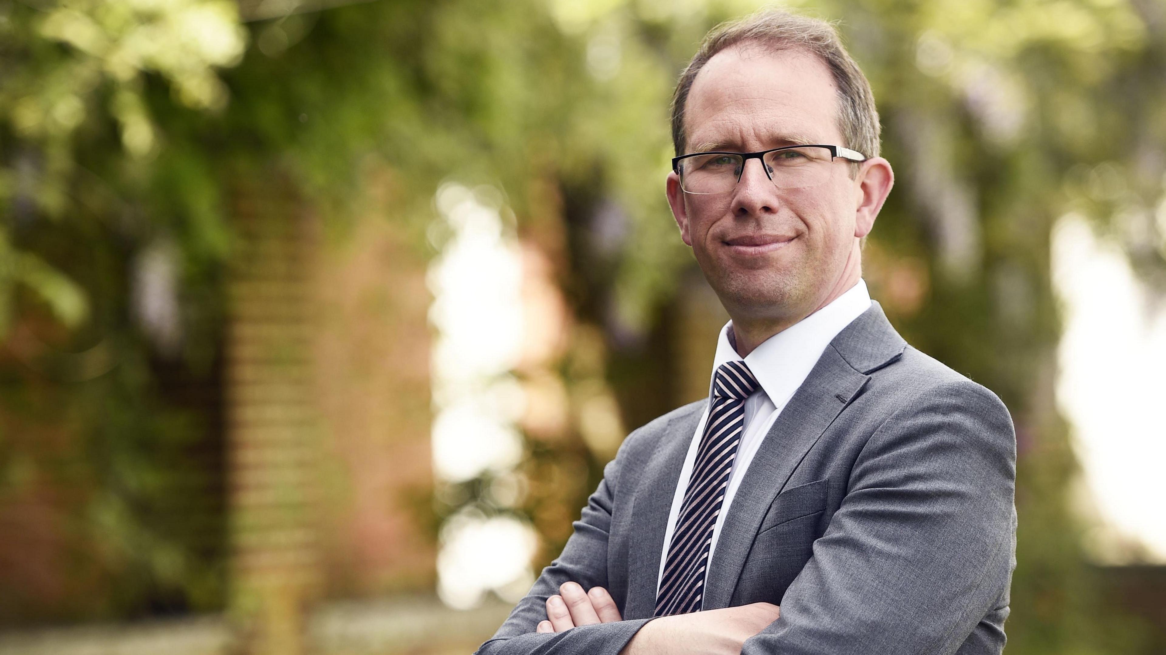 Matthew Barber, who has short brown hair and glasses and is wearing a grey suit, with his arms folded