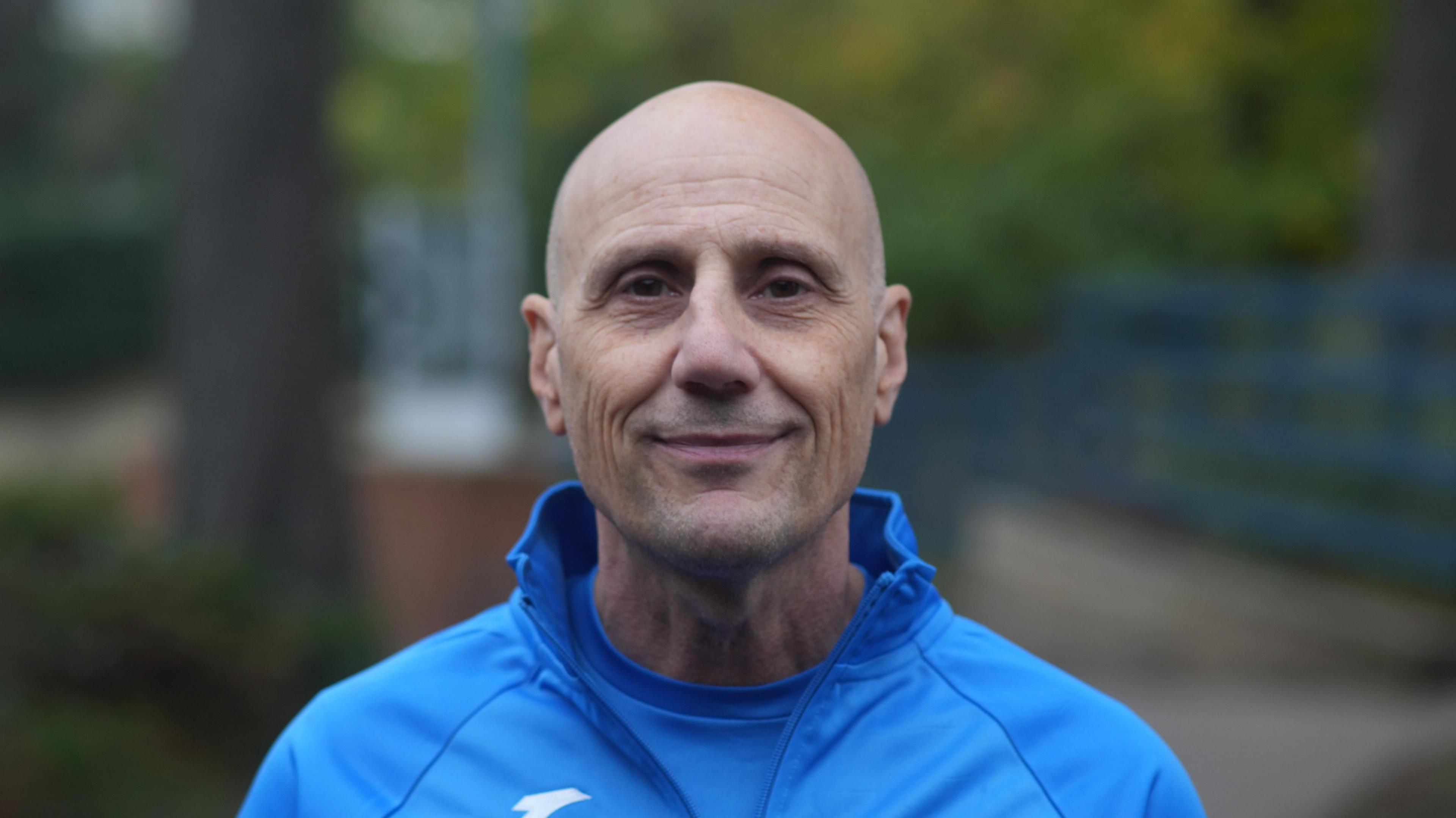 Nino Severino smiles at the camera. He is bald and wearing a blue jumper with a blue top underneath.