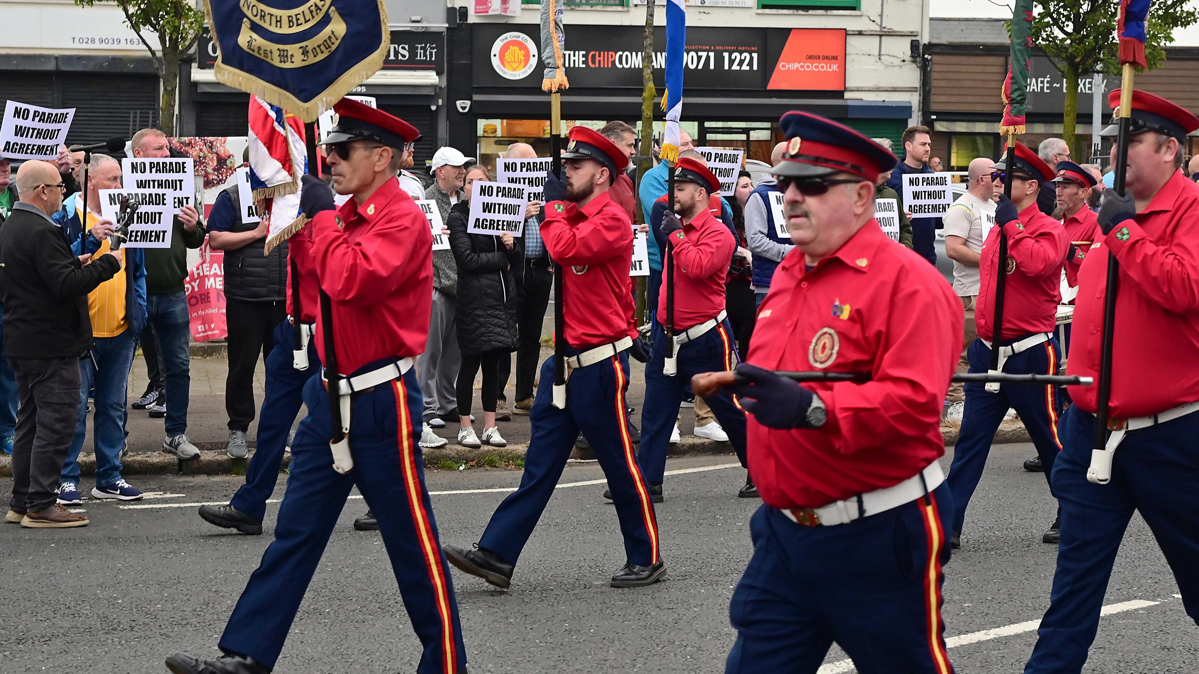 Band wearing red uniform marches past shops where protesters hold banners aloft