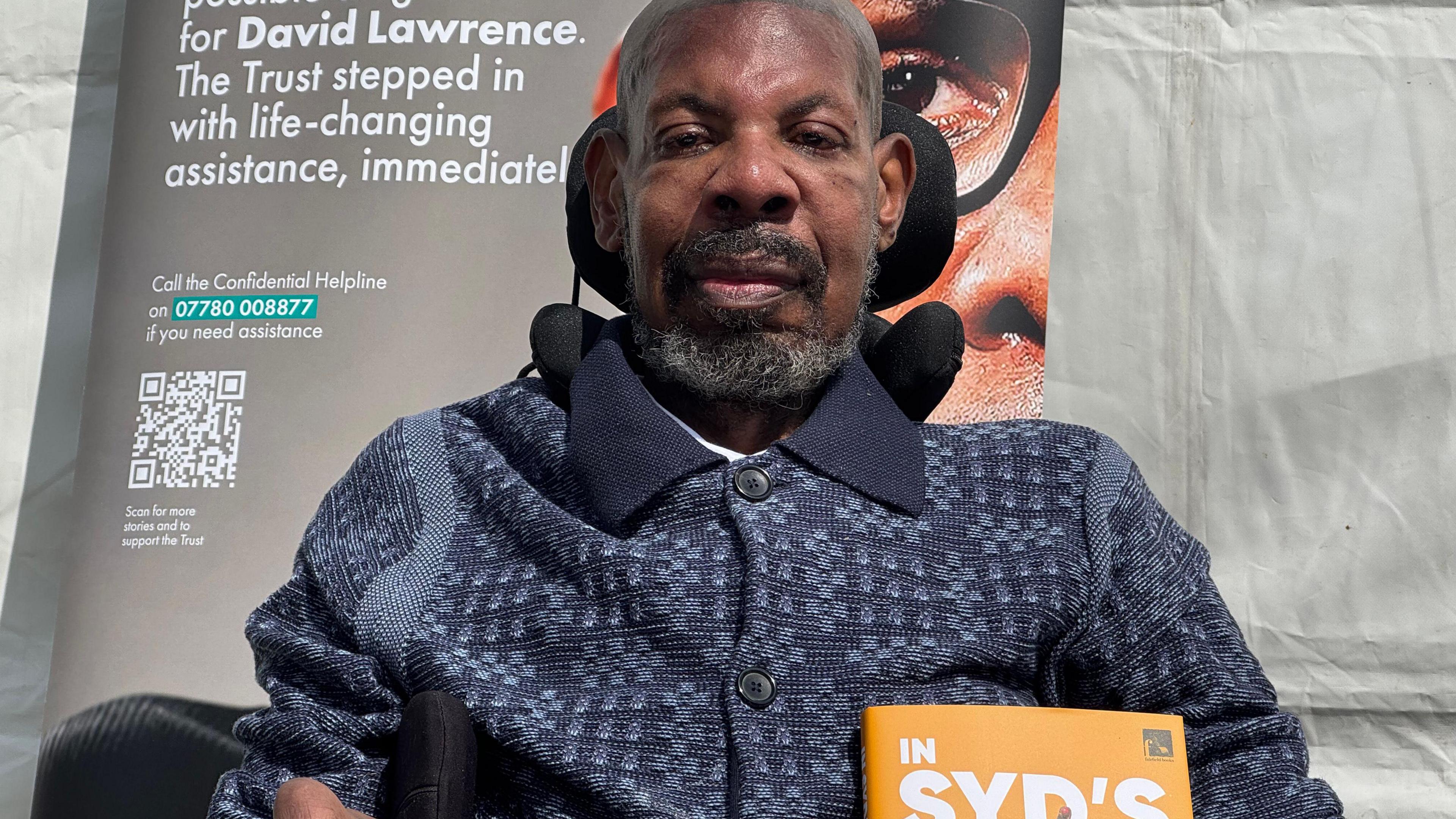 Syd sitting in a wheelchair with a dark polo shirt on, short beard and hair, smiling at the camera. There is a poster behind him and he is outside by a white tent.
