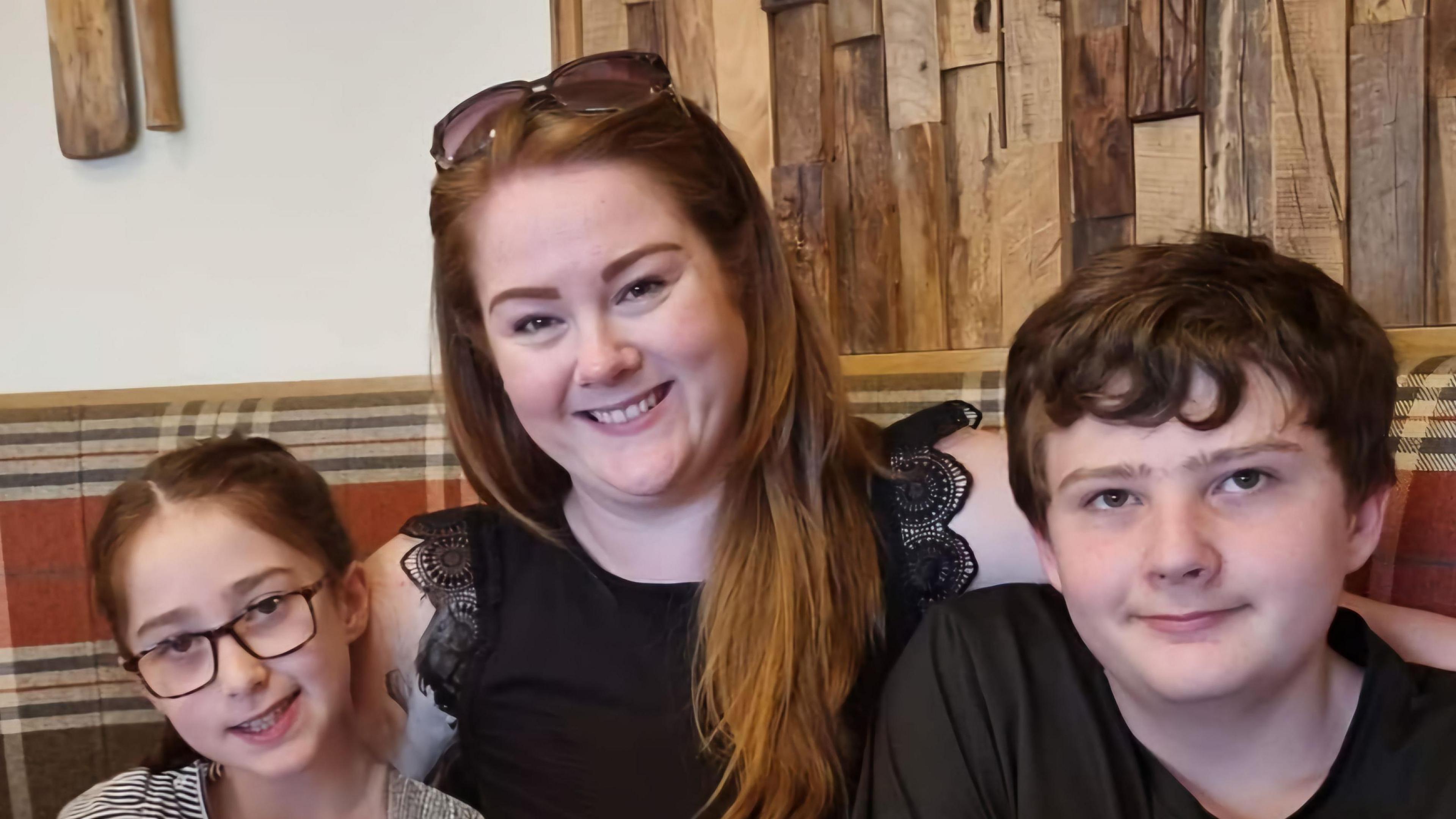 A woman with long brown hair with sunglasses on her head and wearing a black sleveless top sits between two children a girl on the left with black framed glasses and a boy on the right with dark hair and a black t-shirt on