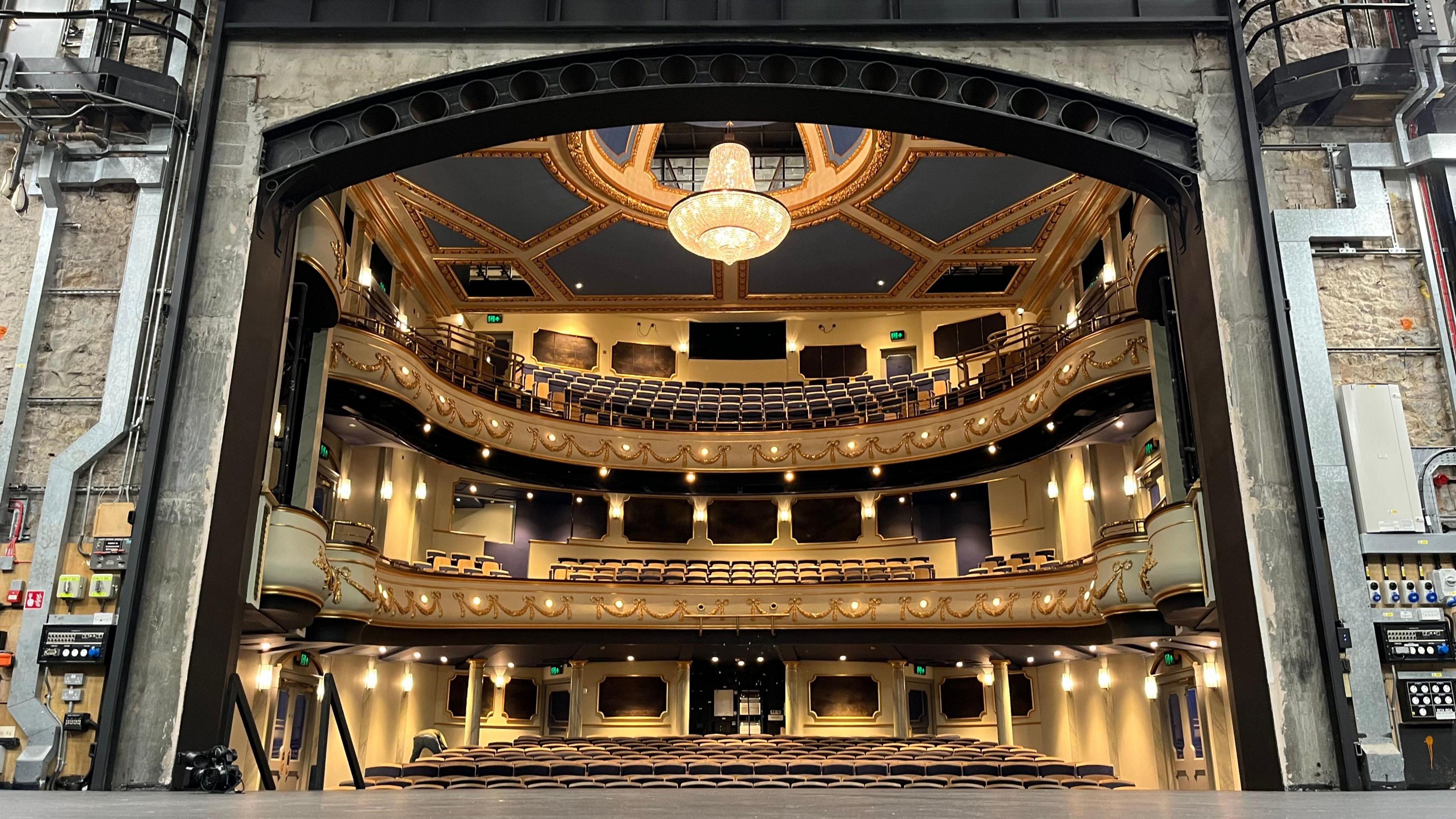 The opera house viewed from the stage. It has three tiers of seats and the colours used are gold and purple. There is a large ornate chandelier in the centre of the ceiling. To the left and right of the stage are boxes of switches.