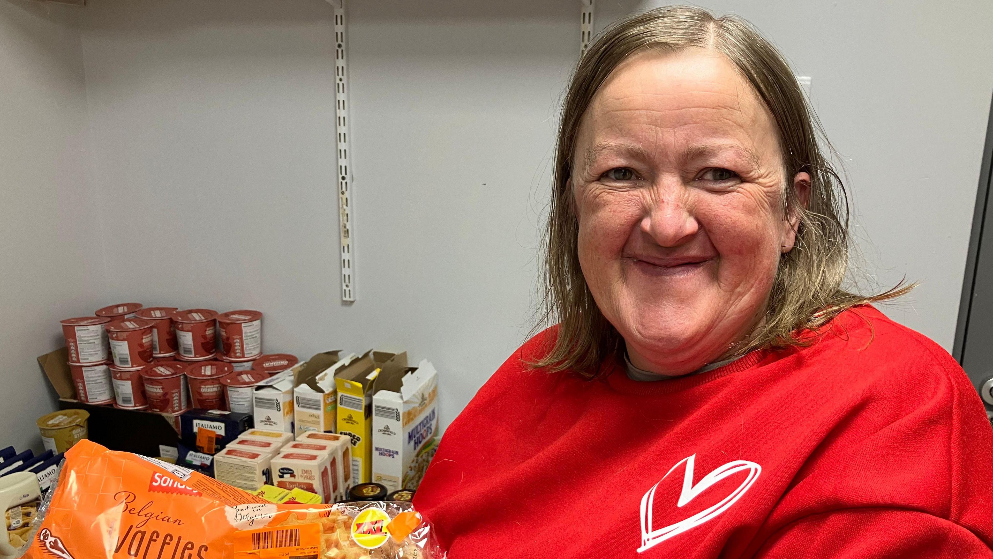 A happy looking woman in a red top with the symbol of a heart printed on it stands smiling into the camera.  She is holding up a bag of Belgian waffle.  It is just one of the items she has picked up.  In the background are a row of ready made porridge in pots stacked in a row.