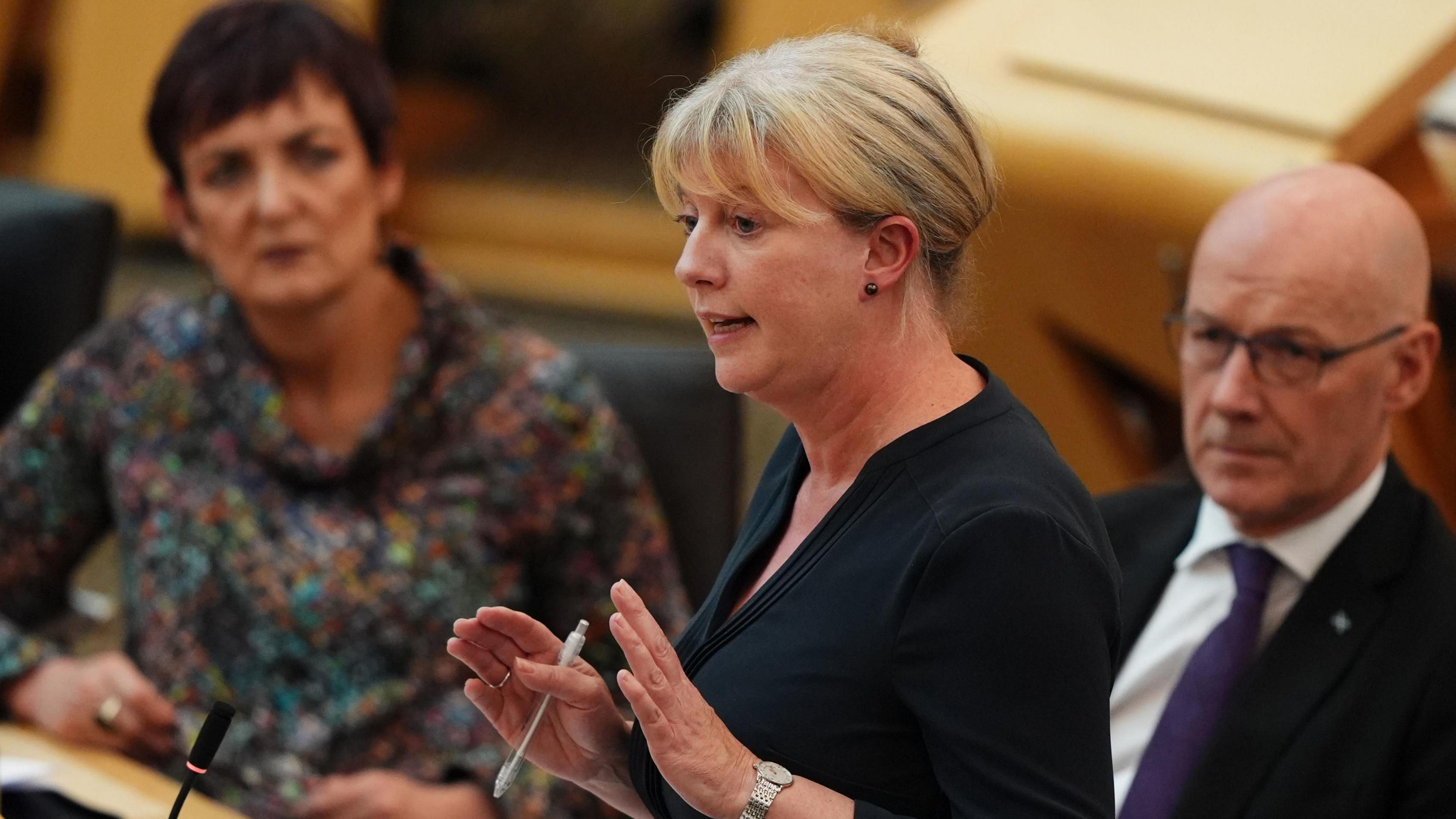 Shona Robison standing speaking in the Scottish Parliament. She is in the foreground, with her hands apart, holding a pen. Her hair is tied back and she is dressed in black. Seated behind her are Angela Constance, to the left, and John Swinney.