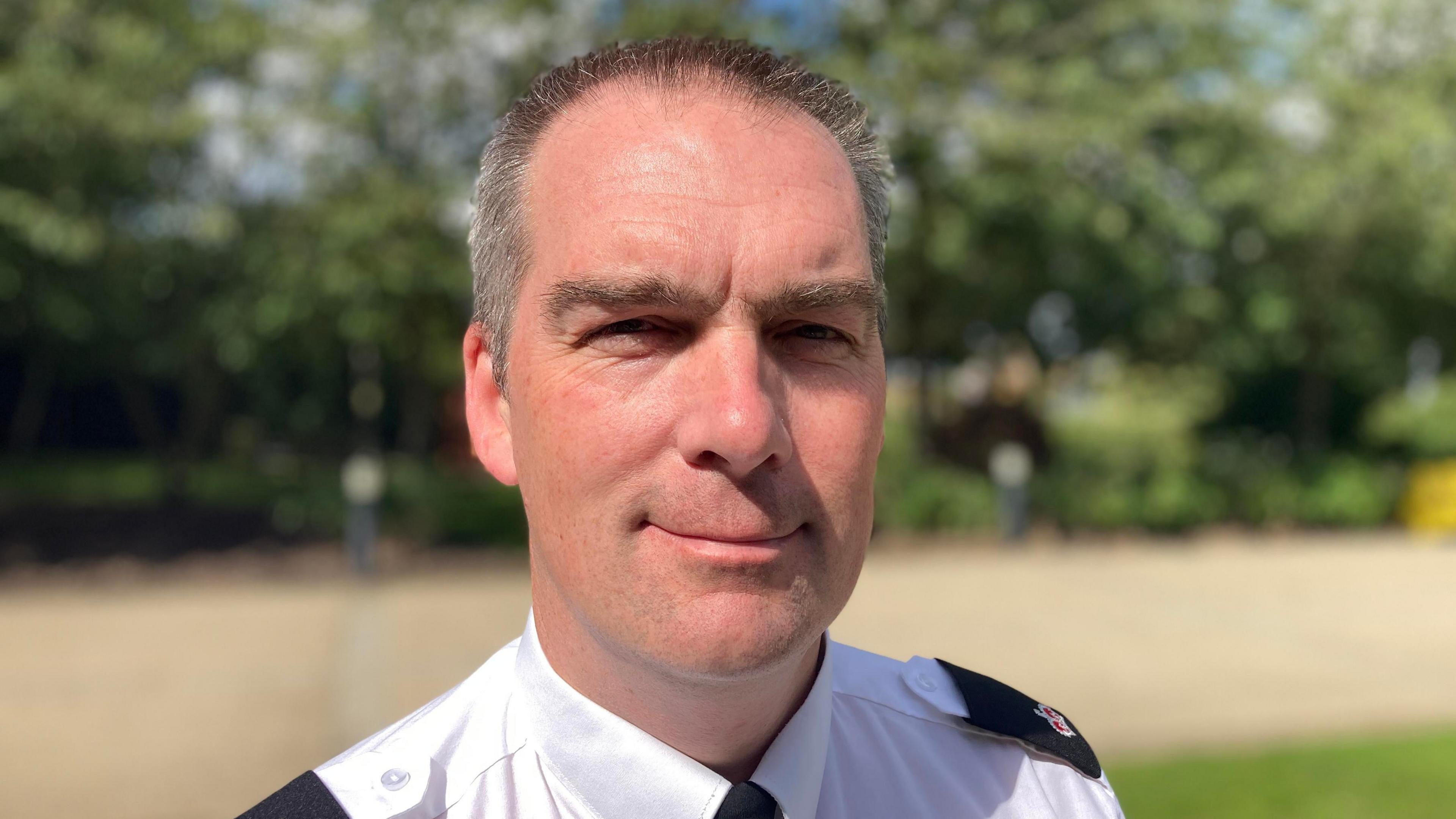 Det Supt Gray with short dark hair, smiling at the camera and wearing a white police shirt with black tie and epaulettes. There is a hedge behind him. 