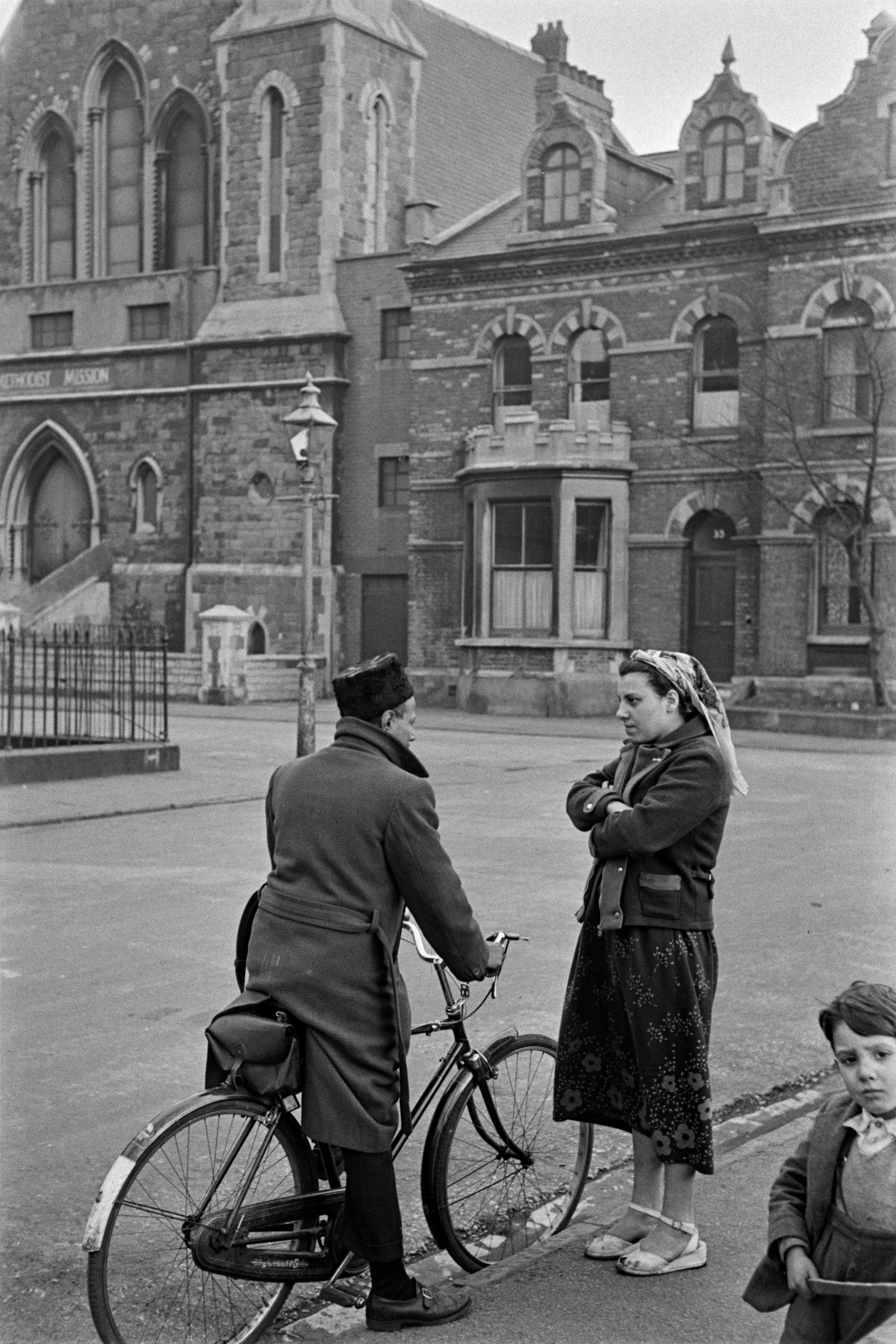 A man and a woman in conversation on a street. The man is on a bike.