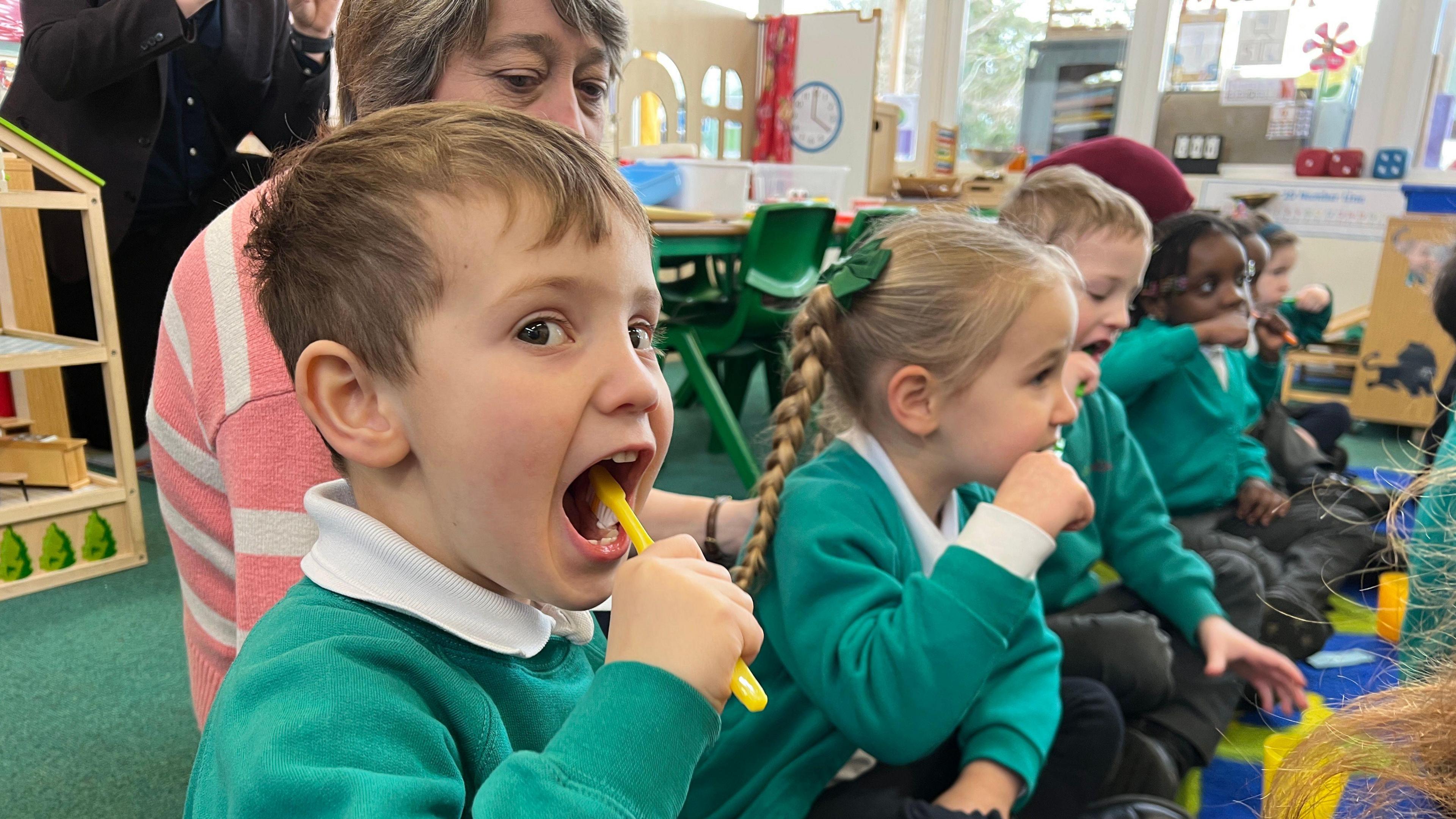 A child in a school classroom with a yellow toothbrush brushes his teeth whilst looking at the camera. He is wearing a teal-coloured school uniform, as are the 4 students in the background who are all also brushing their teeth. A woman looks over the top of the nearest child wearing a pink and grey striped cardigan.