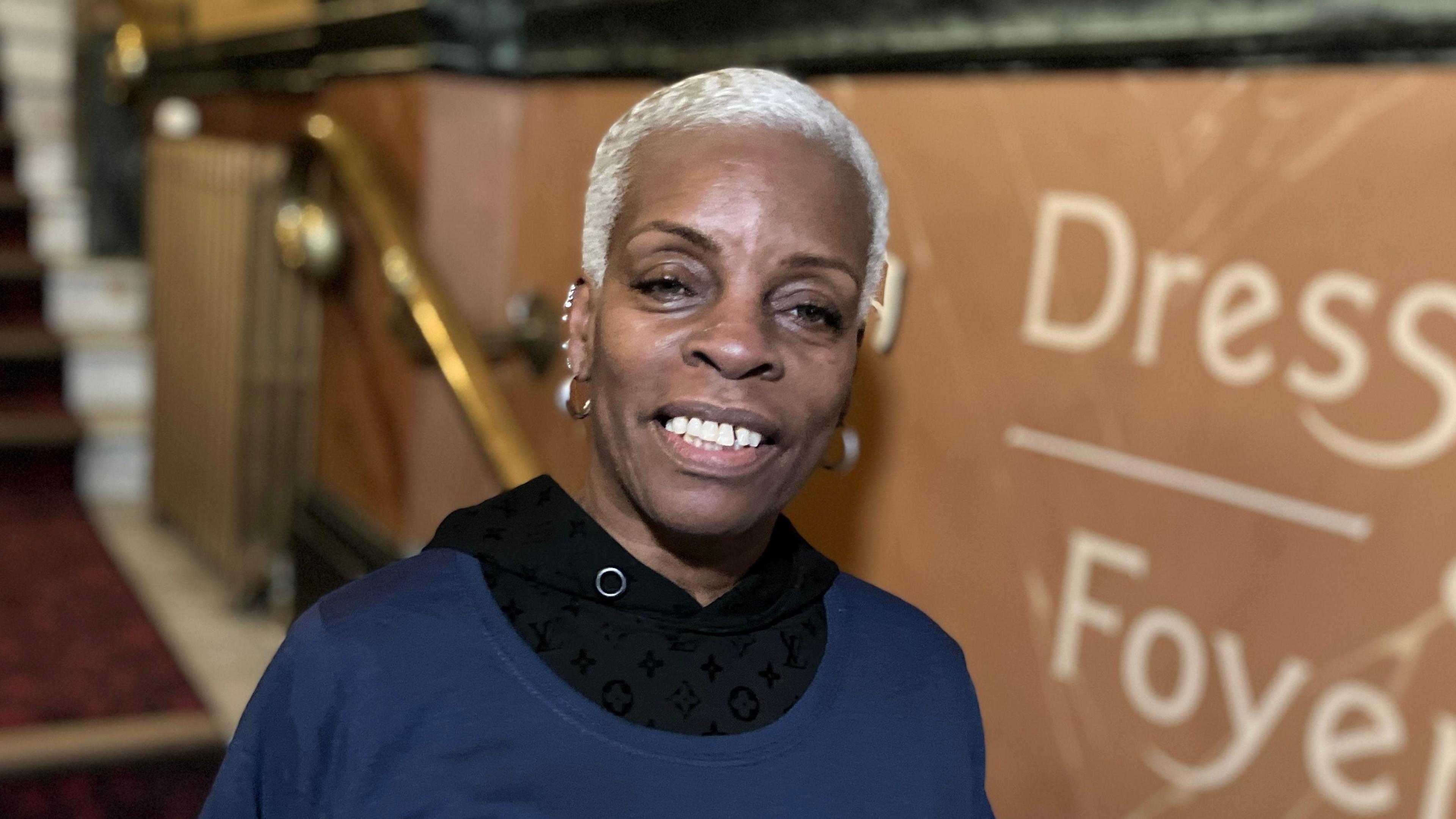 Janette Collins MBE is smiling at the camera. A black woman with short blonde hair, she is wearing a navy blue jumper with a black top underneath. She is pictured in the foyer of Hackney Empire