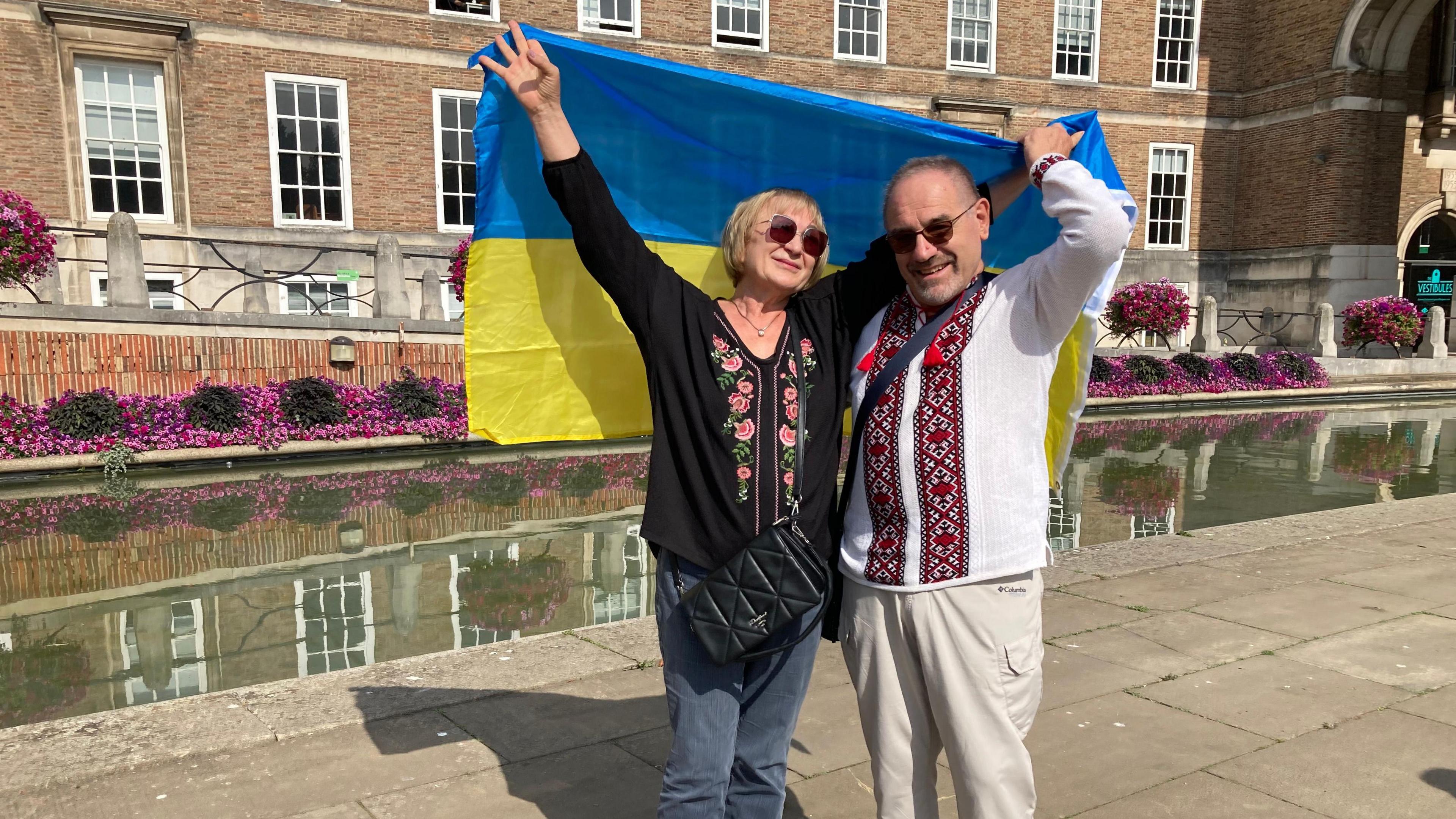 A woman and a man arm in arm, holding up a blue and yellow Ukrainian flag behind them as they stand on Bristol's College Green. They are both wearing Ukrainian-style embroidered clothes.