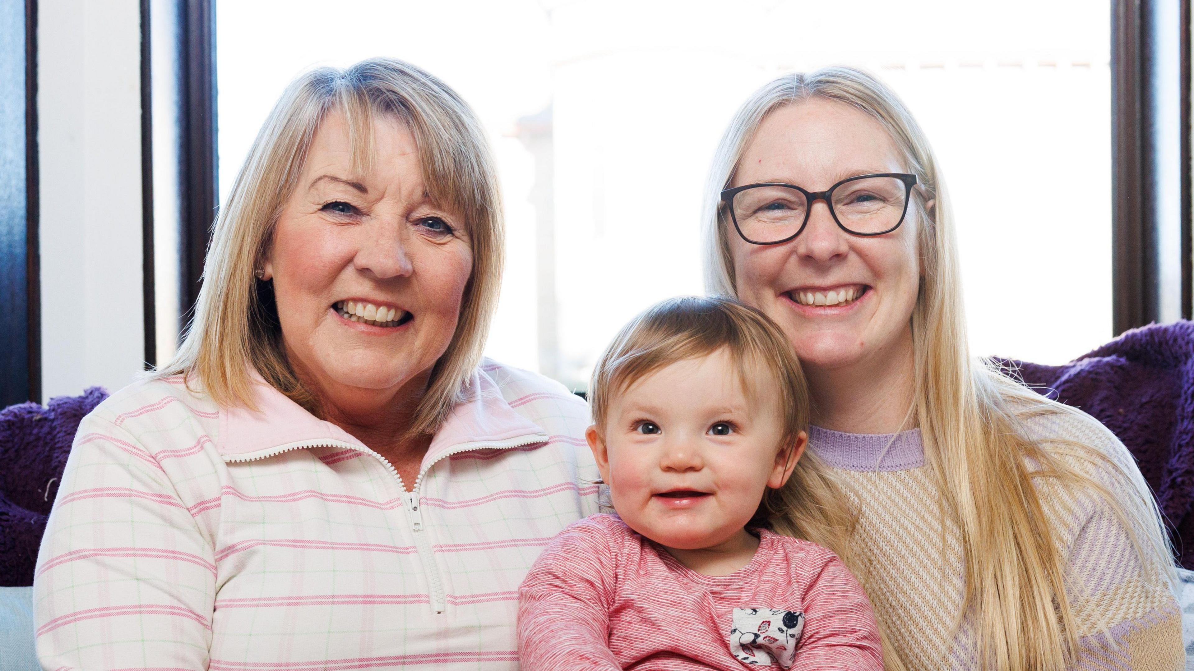 A 61-year-old woman wearing a pink and white striped top and a 35-year-old woman with a yellow and purple striped top and glasses, hold a 10-month-old girl, who is wearing a pink top. They are all sitting on a settee with a window behind them.