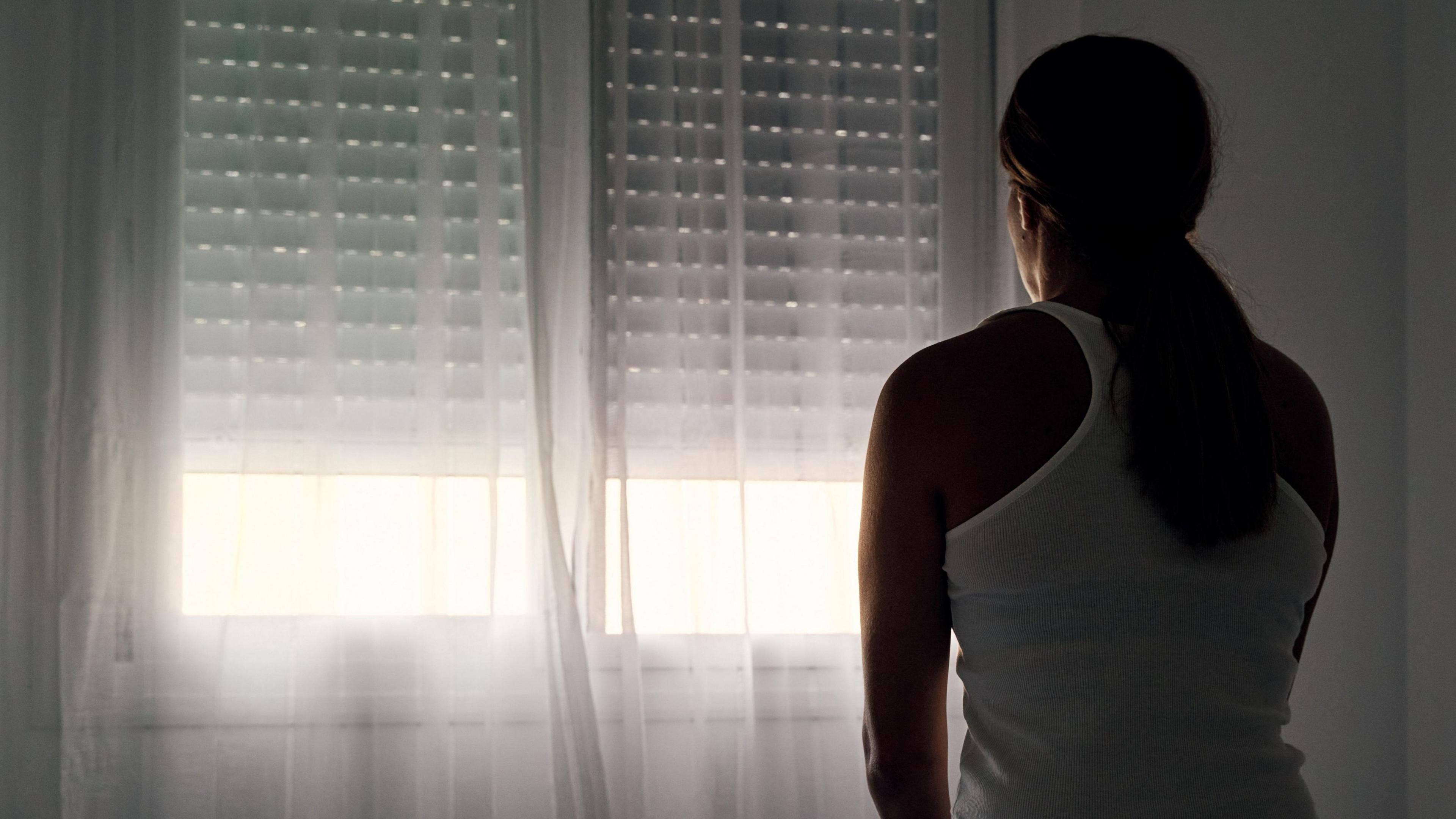 A woman sits on her bed facing the window. The blinds are nearly all the way down. Her hair is tied back and she's wearing a white vest top.