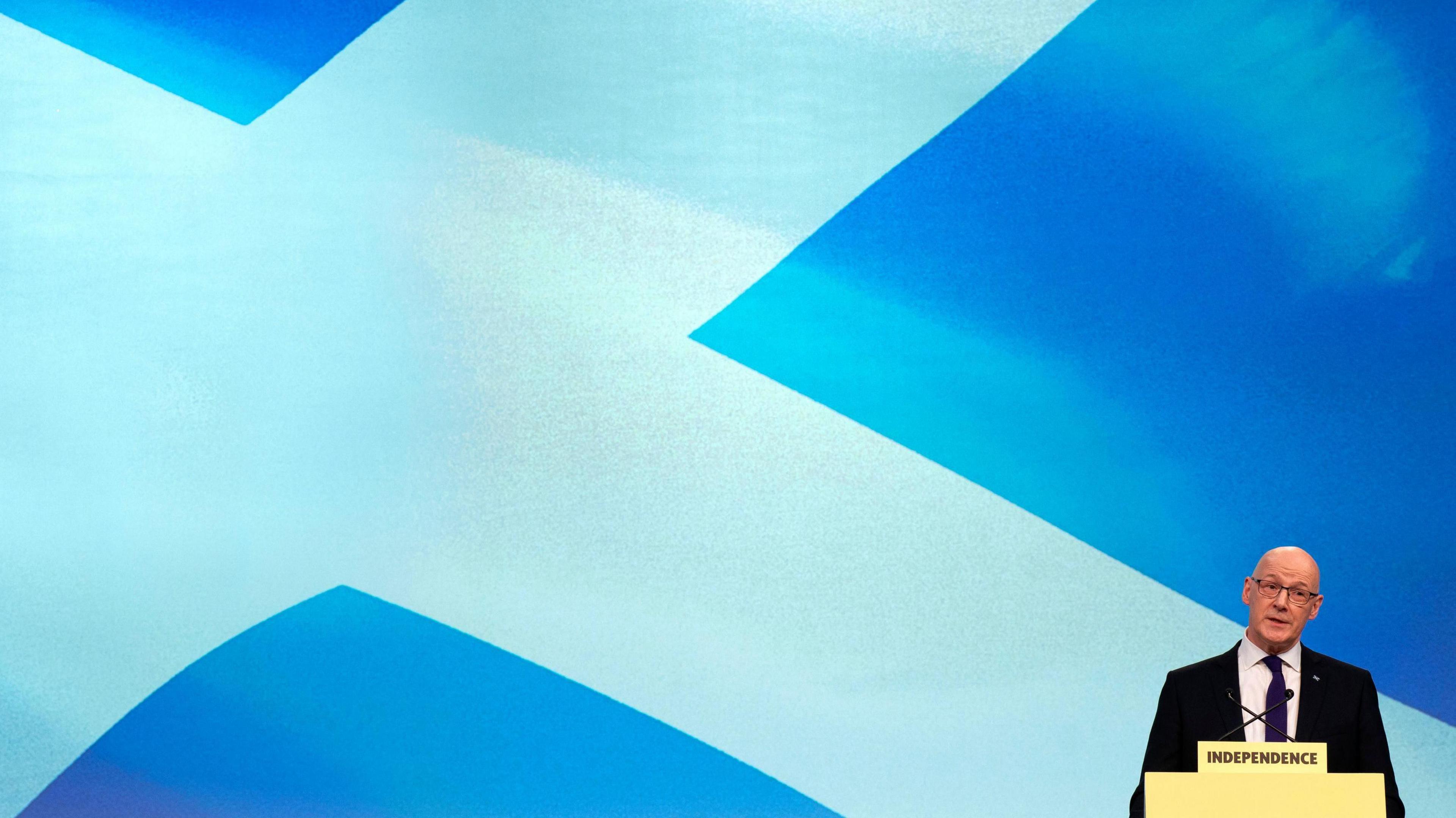 John Swinney stands behind a lectern on the bottom right of the image, with a large saltire on a screen behind him.