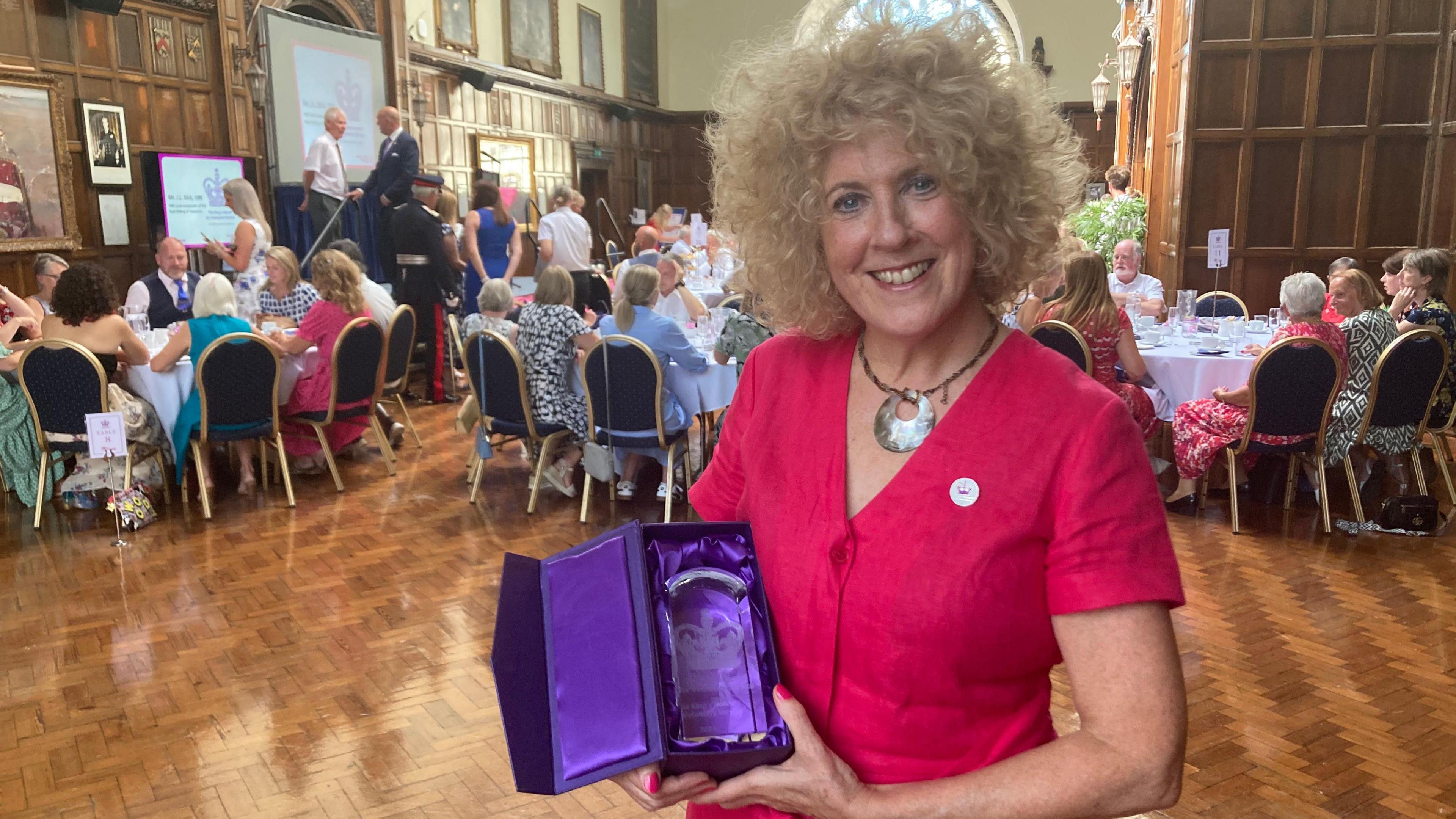 Ms Barlow wears a bright fusia dress and has blond curly hair in a bob. She holds the glass award which sits in a purple satin box. The volunteers sit around tables in the background within the wood panelled room of the Guildhall.