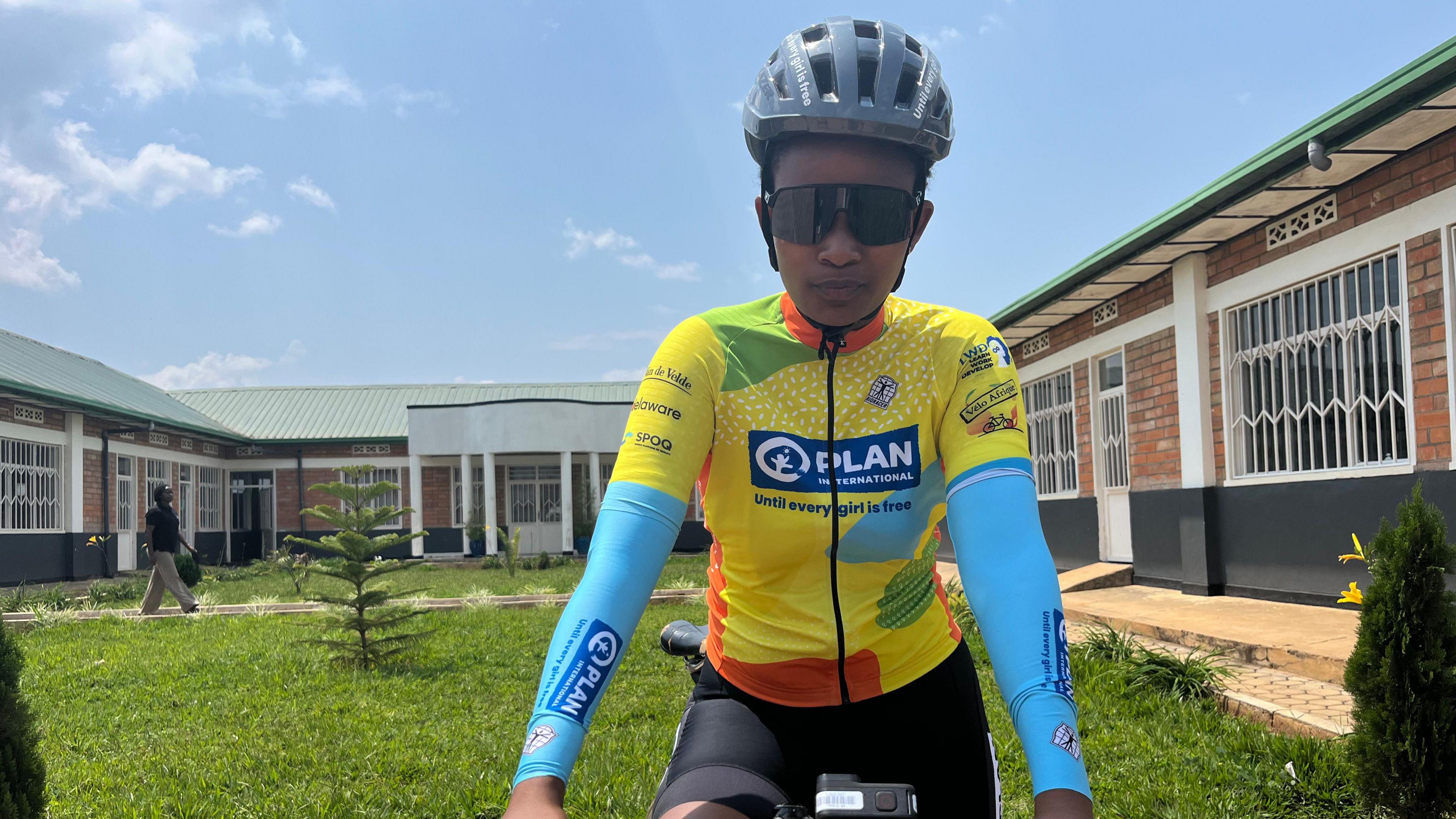 Olivia Manigragena wears a grey helmet, sunglasses and a brightly-coloured, predominantly yellow, cycling jersey as she sits on a bicycle and looks into the camera ahead of a race at the Bikes for Future centre in Bugesera. Three sides of a single-storey red brick building with white windows and a green roof can be seen in the near background behind her surrounding a patch of green grass