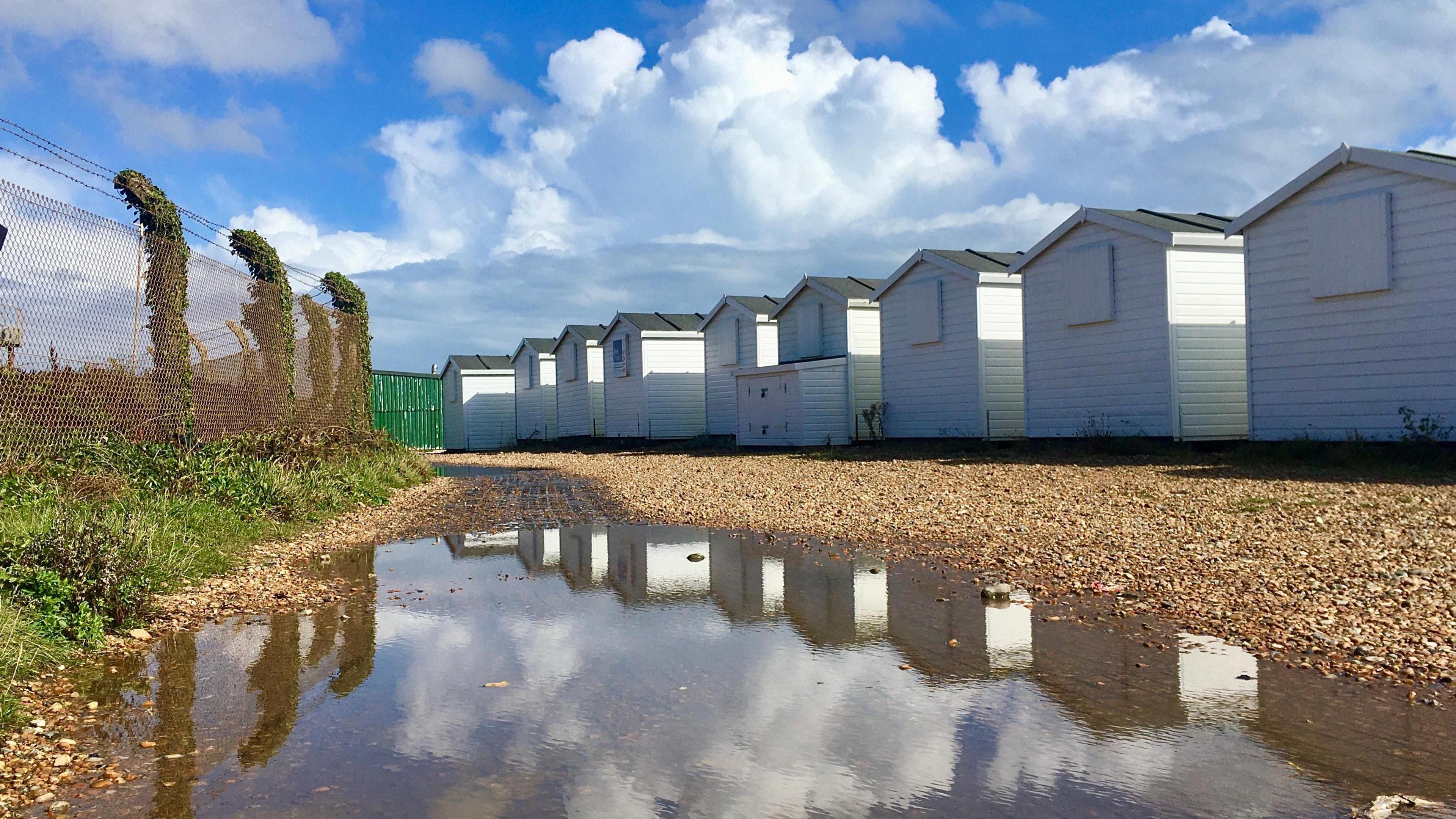 A row of beach huts with pebbles in front and a large puddle in the foreground, with a blue sky and white clouds overhead